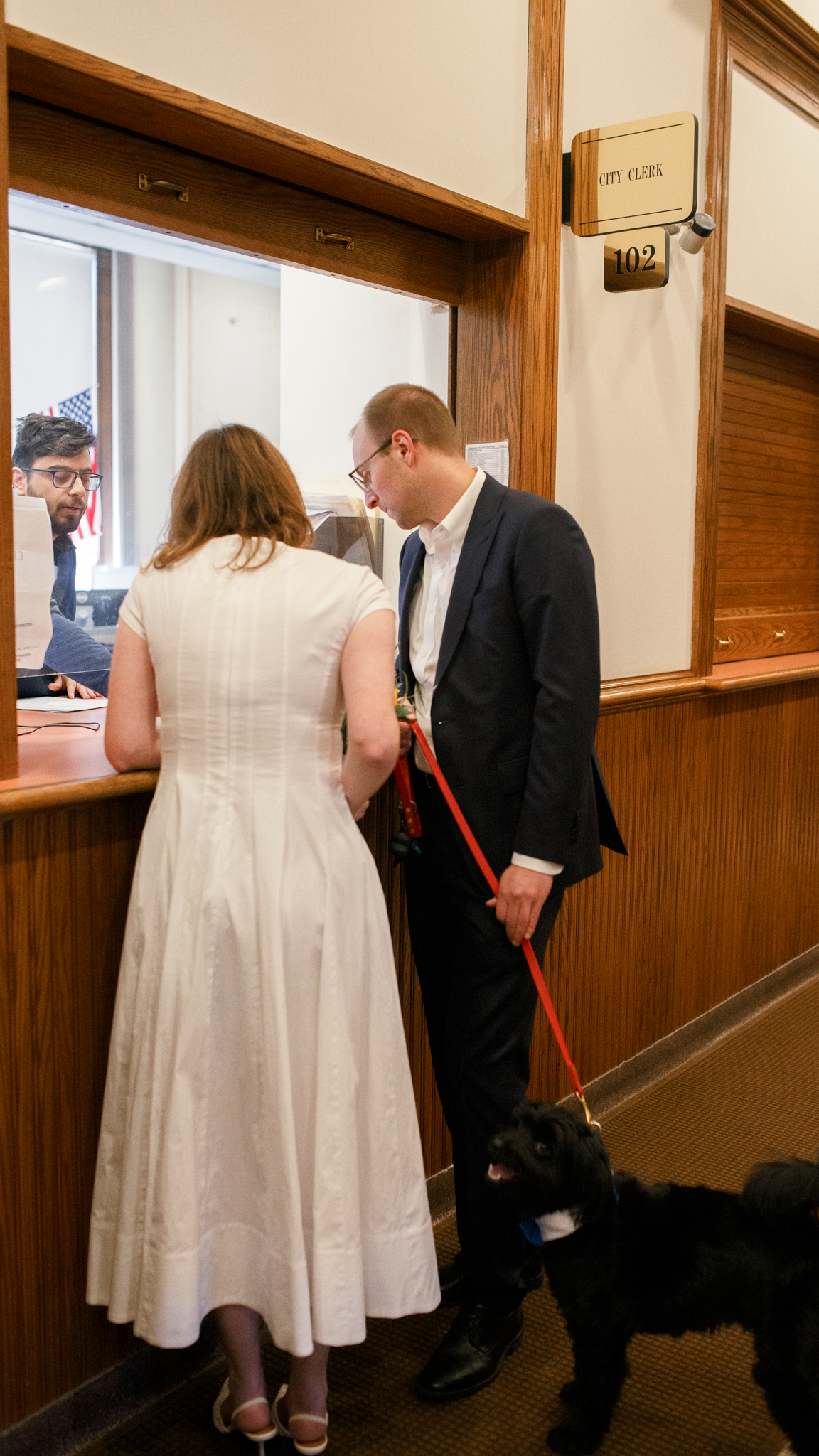 People in formal attire wait in line at an office counter with wood paneling and numbered windows.