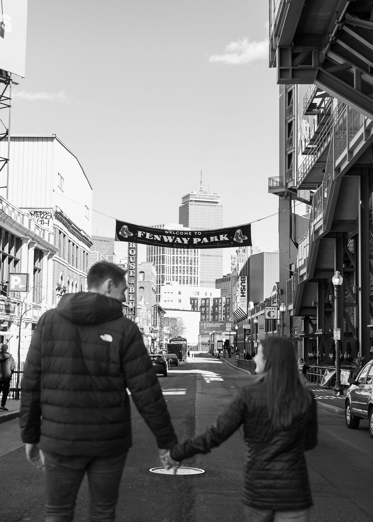 Black and white engagement photography of a couple walking toward the Fenway Park sign, capturing the urban feel of a Boston proposal.