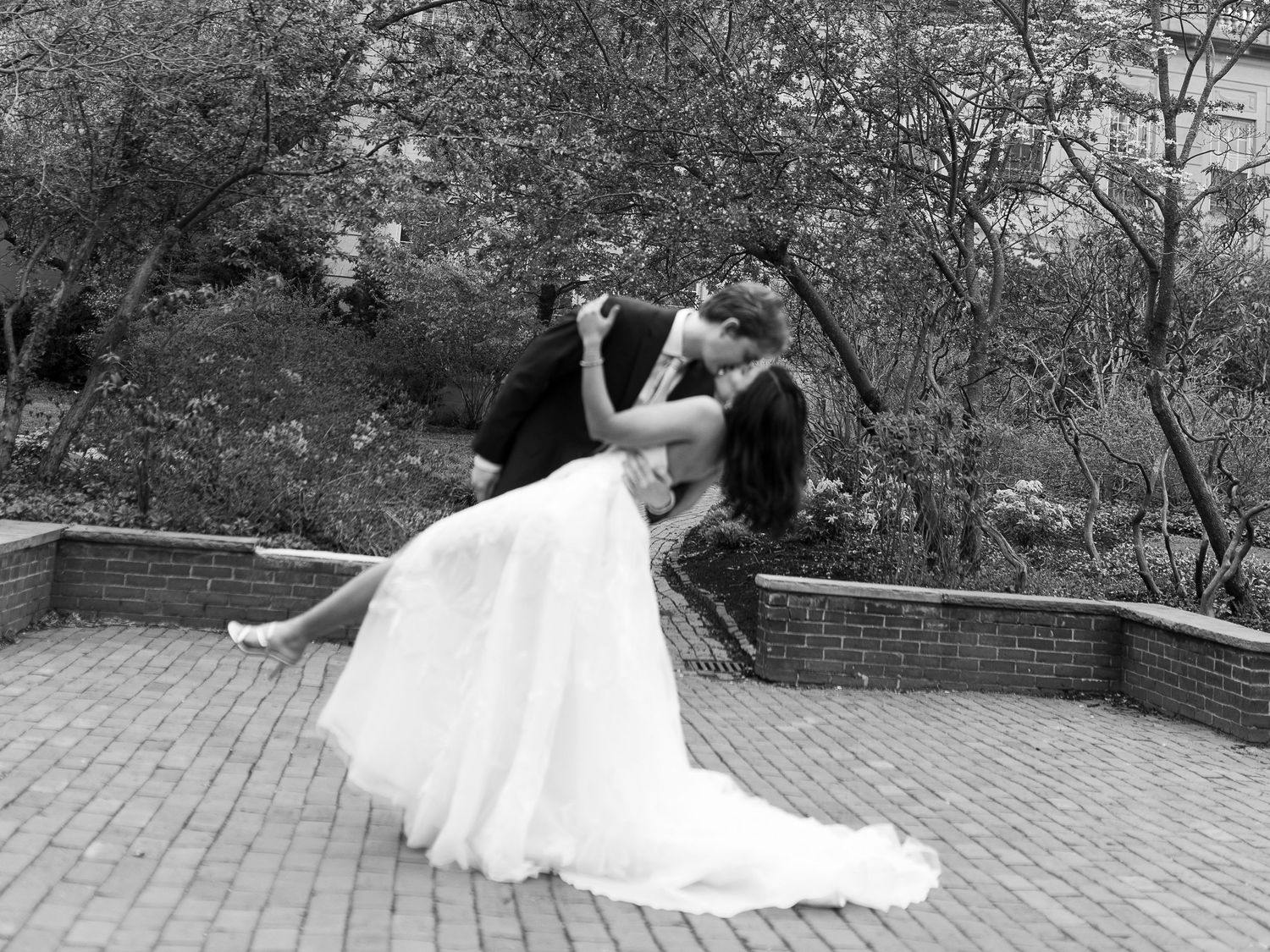 Black and white wedding photo of couple sharing romantic dip kiss on brick terrace surrounded by lush trees.