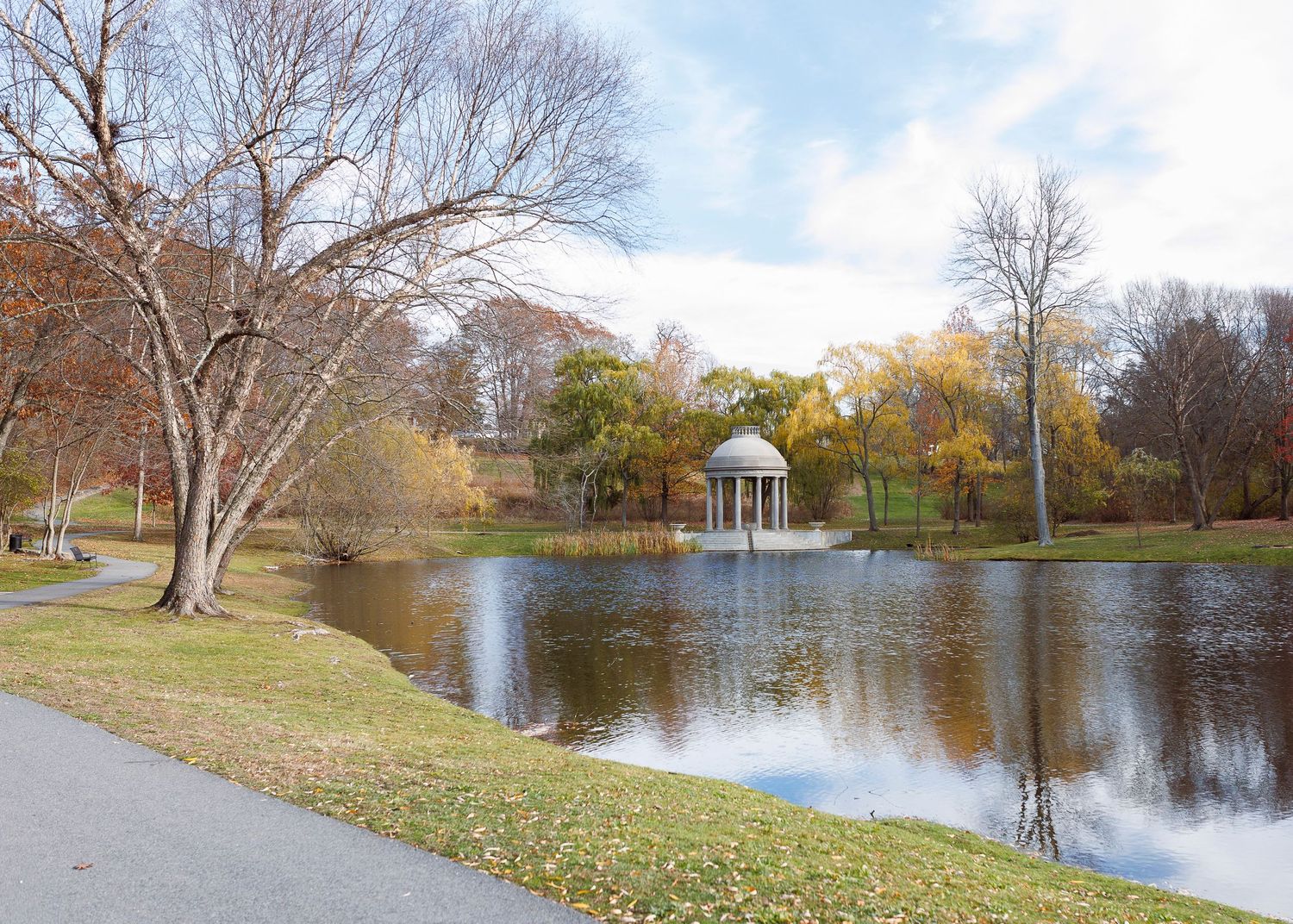 A panoramic view of a serene pond with a fountain and bare trees along the grassy shoreline on a sunny autumn day.