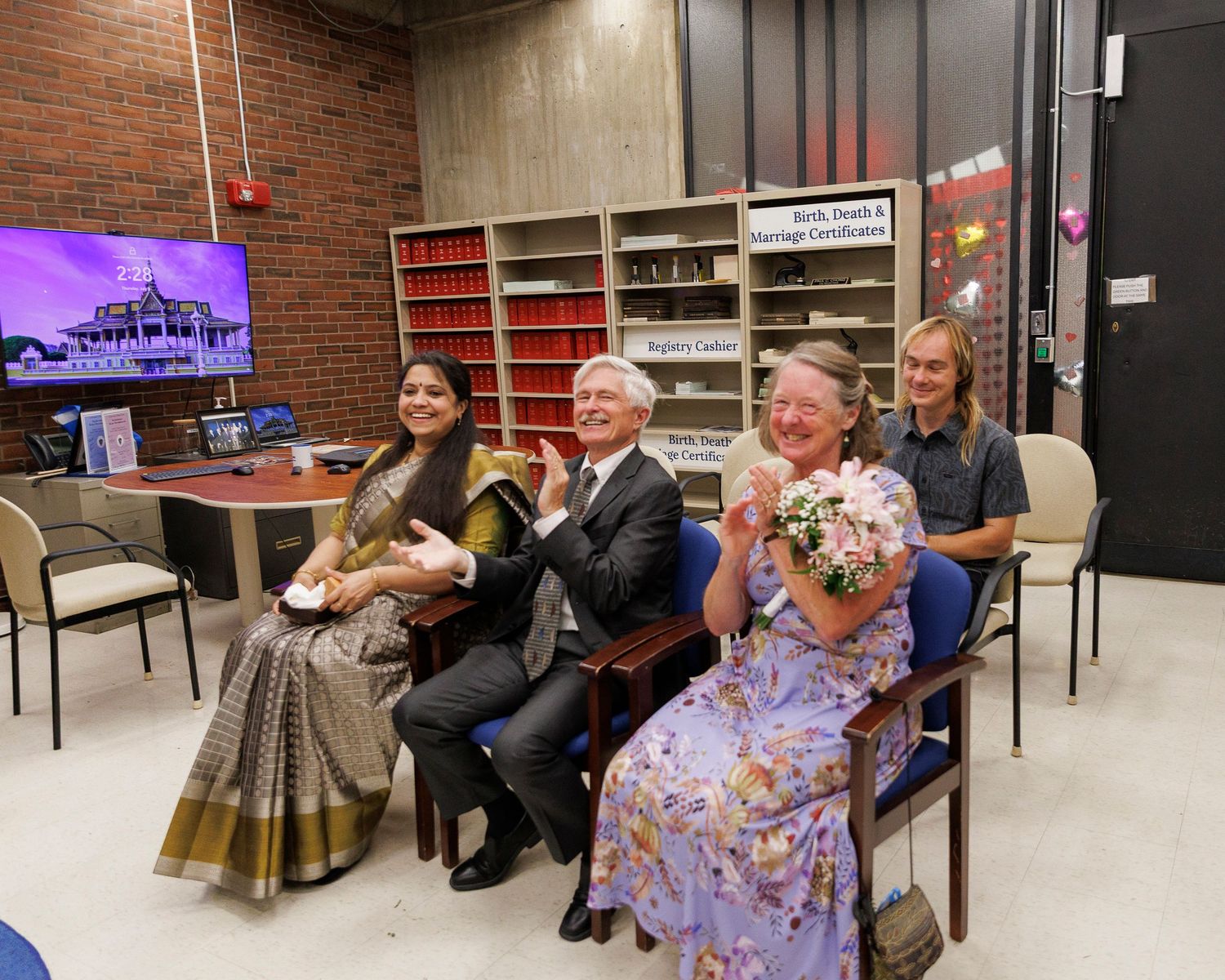 Four people smile and clap together in a decorated room, with a woman in a sari and a man in a suit seated centrally.