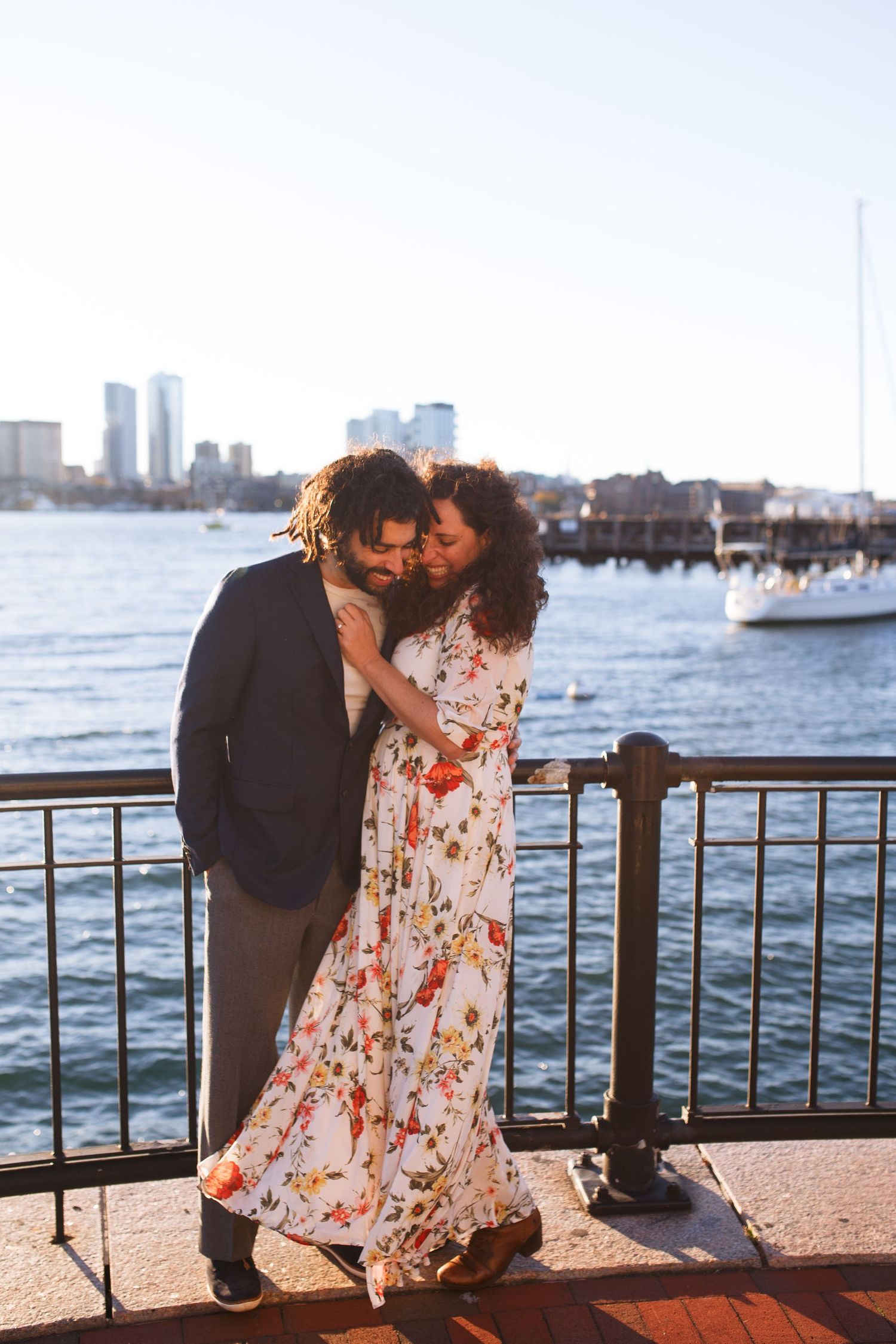 Romantic couple embraces along waterfront railing with city skyline and boats in background at sunset.