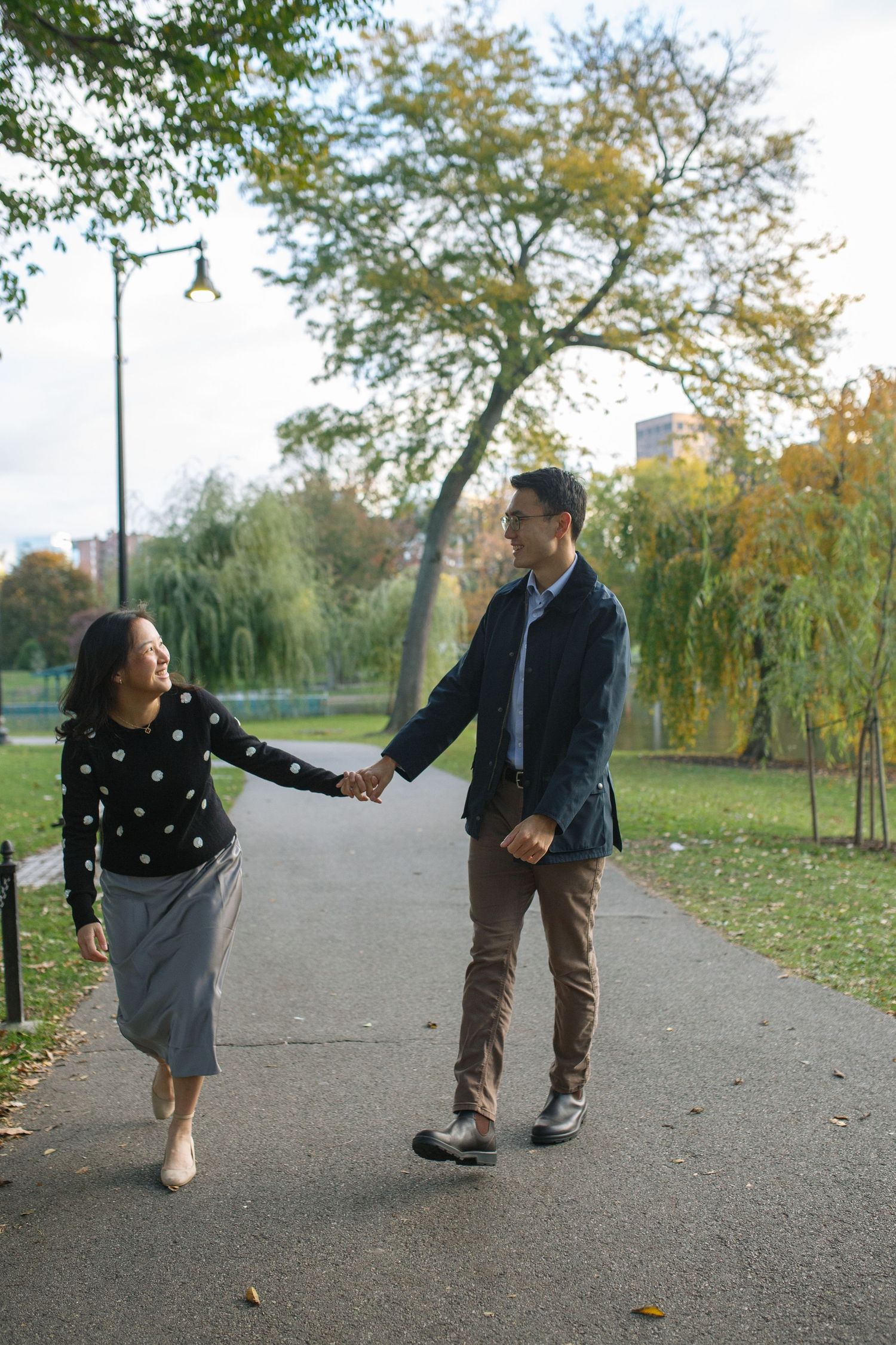 A couple holds hands while walking along a park path surrounded by autumn trees and fallen leaves.