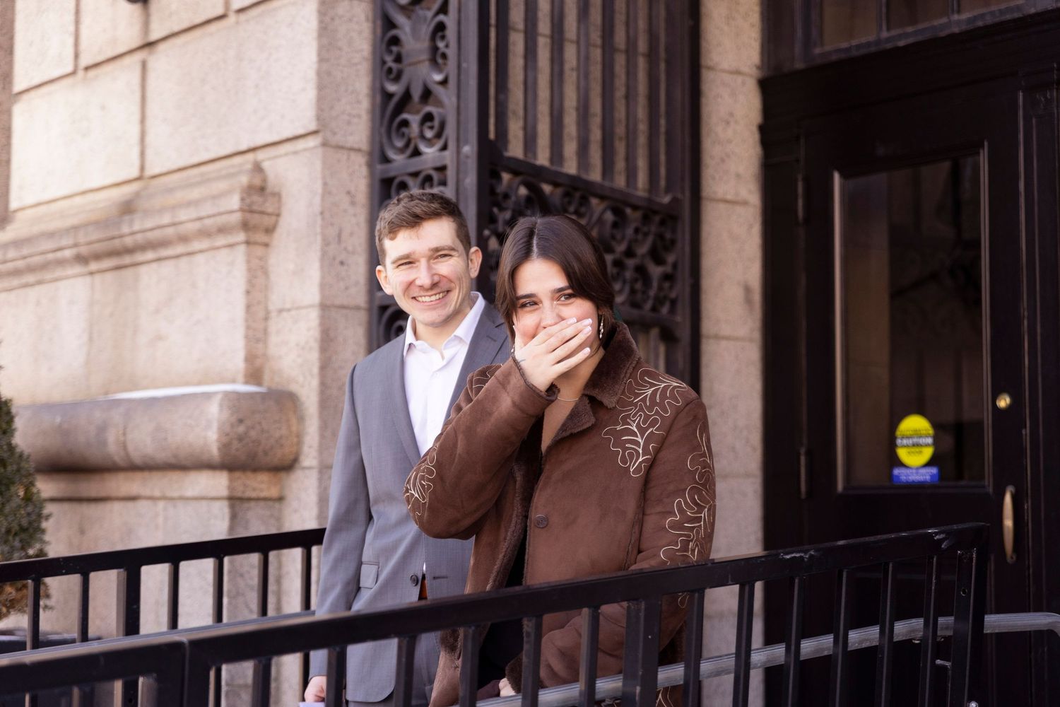 Two people in formal attire stand in front of an ornate stone building with iron gates.