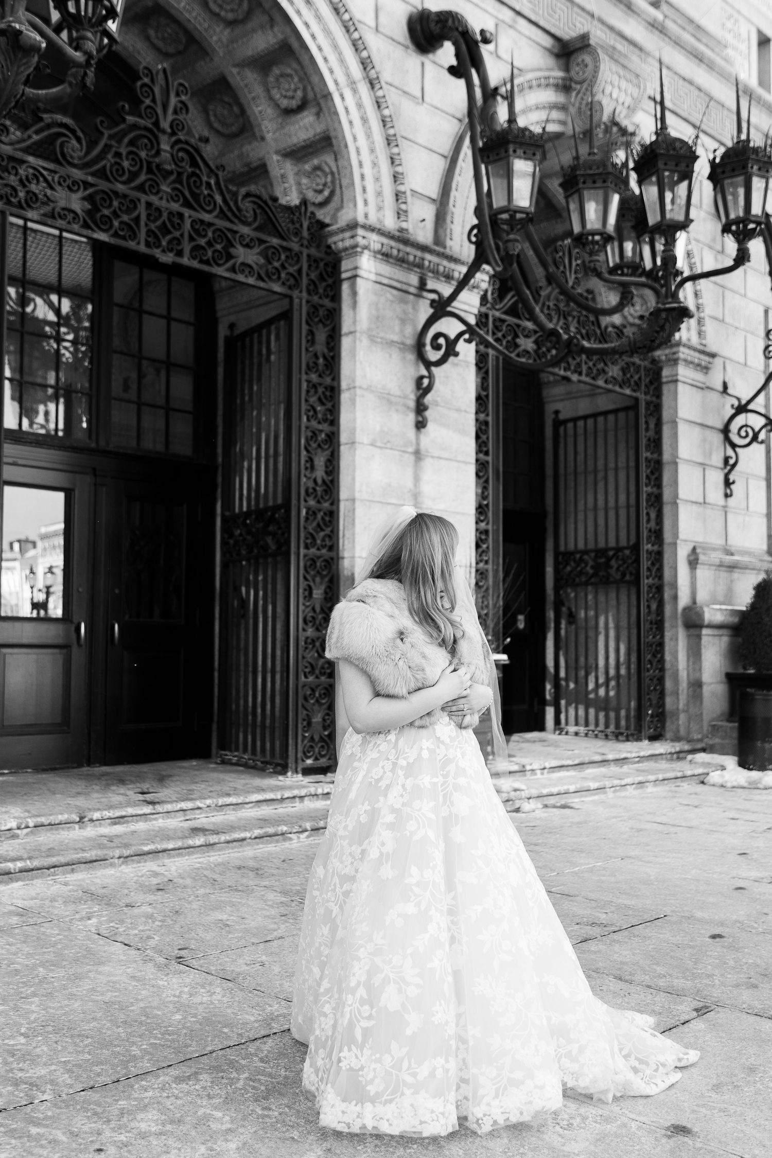 A bride in a white lace gown stands outside an ornate building entrance with decorative iron lanterns.