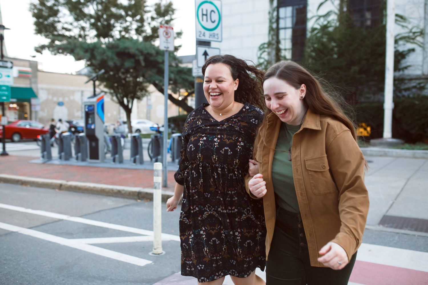 Two people walk and laugh together on a city sidewalk while holding ice cream cones on a sunny day.