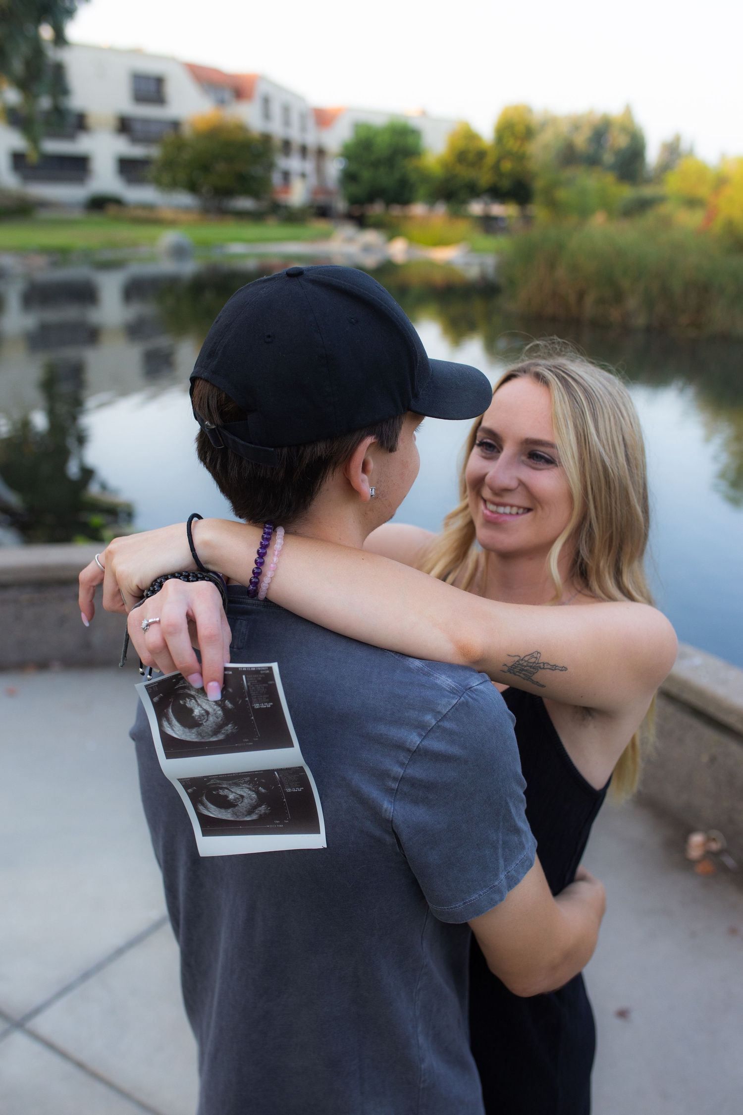 A couple embraces while holding an ultrasound photo by a lake during sunset.