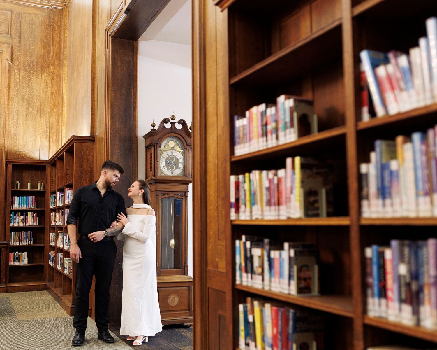 A romantic couple embraces in a wood-paneled library surrounded by tall bookshelves and warm lighting.