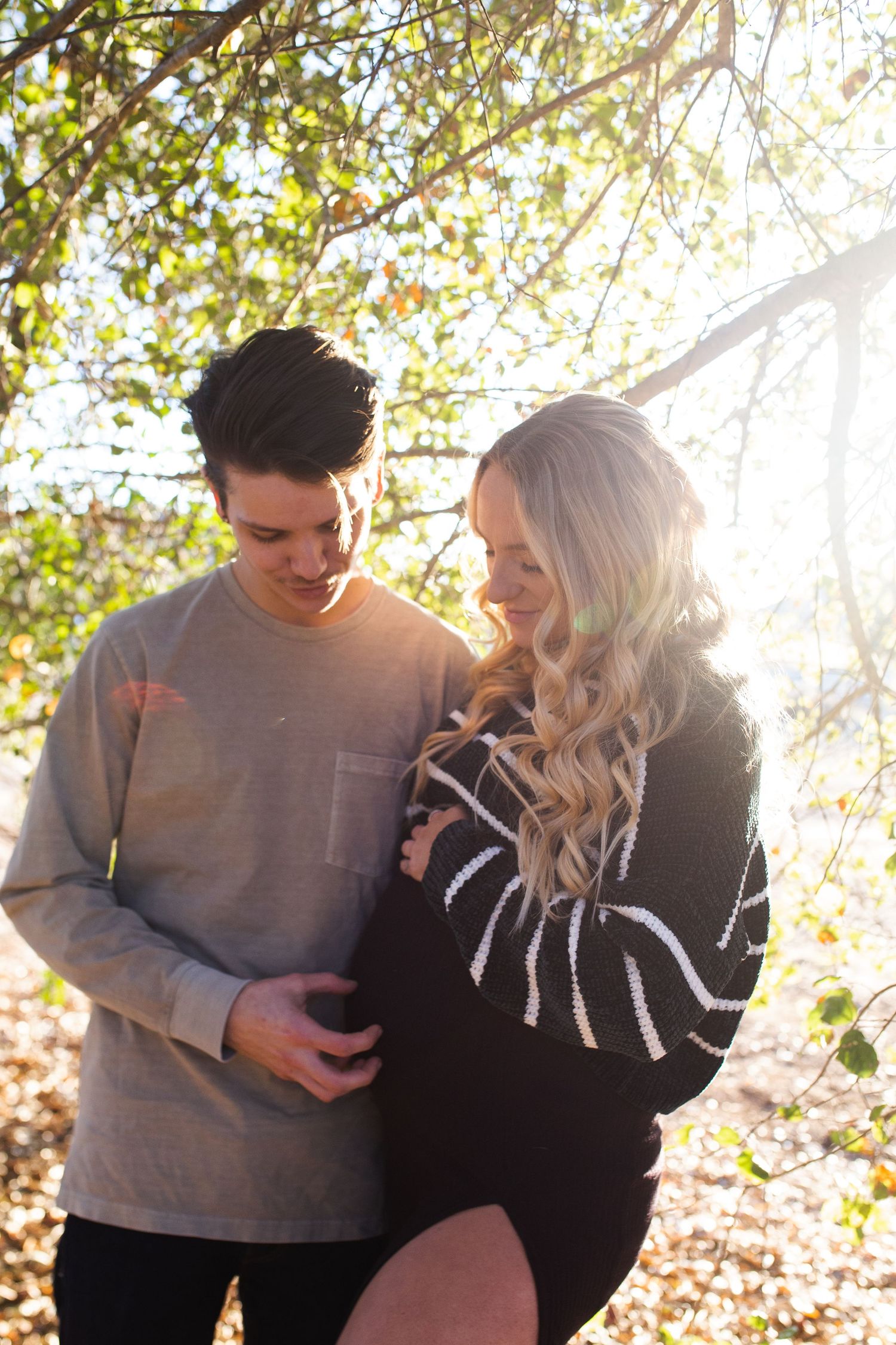 A couple shares an intimate moment together in a sun-dappled outdoor setting with autumn leaves in the background.