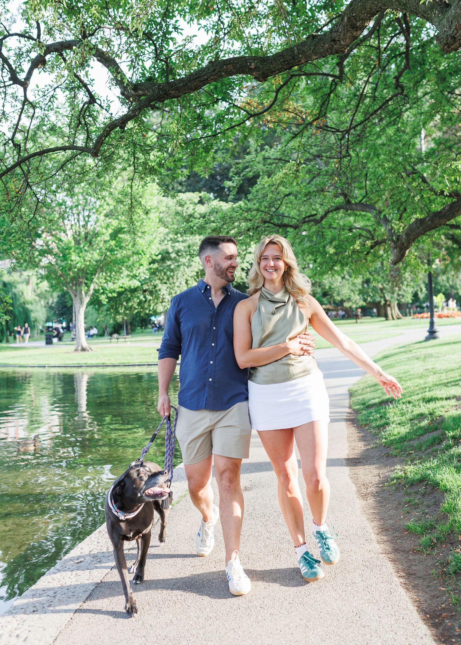 Couple walking their black dog along a scenic park path beside a pond, surrounded by lush green trees on a sunny day.