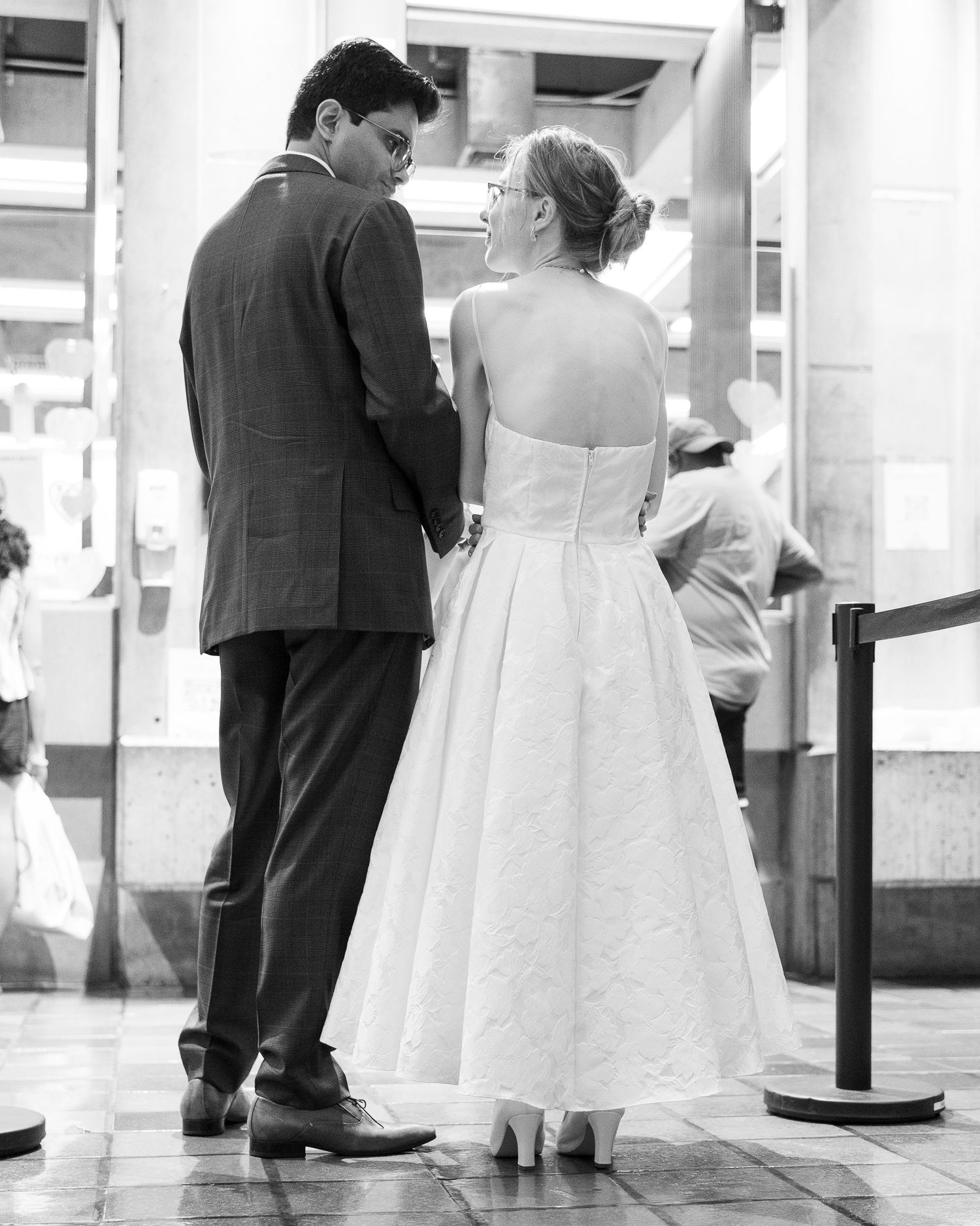 Black and white photo of a couple in wedding attire holding hands from behind in an indoor venue.