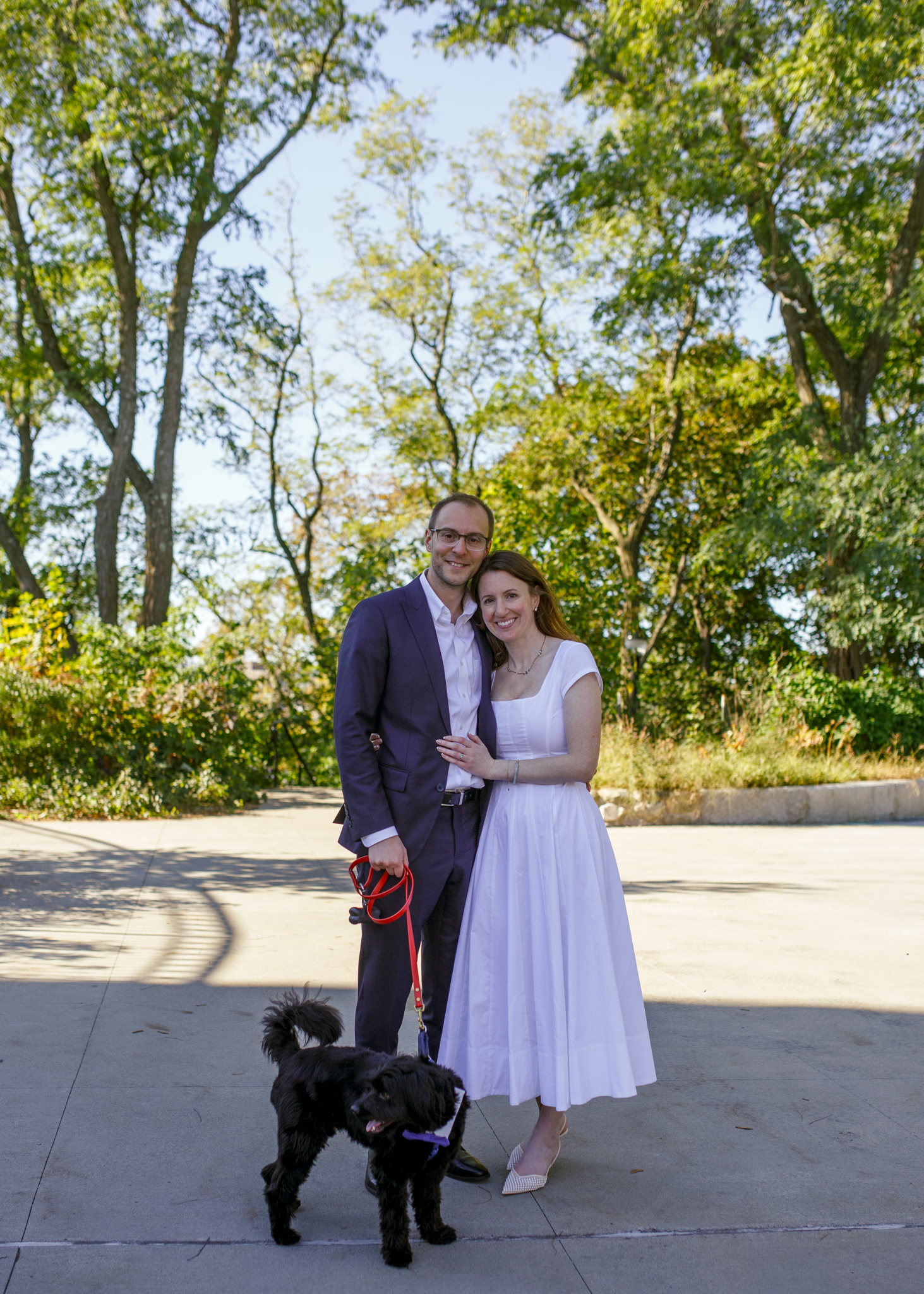 A couple in wedding attire poses with their black dog on a sunny outdoor patio surrounded by trees.
