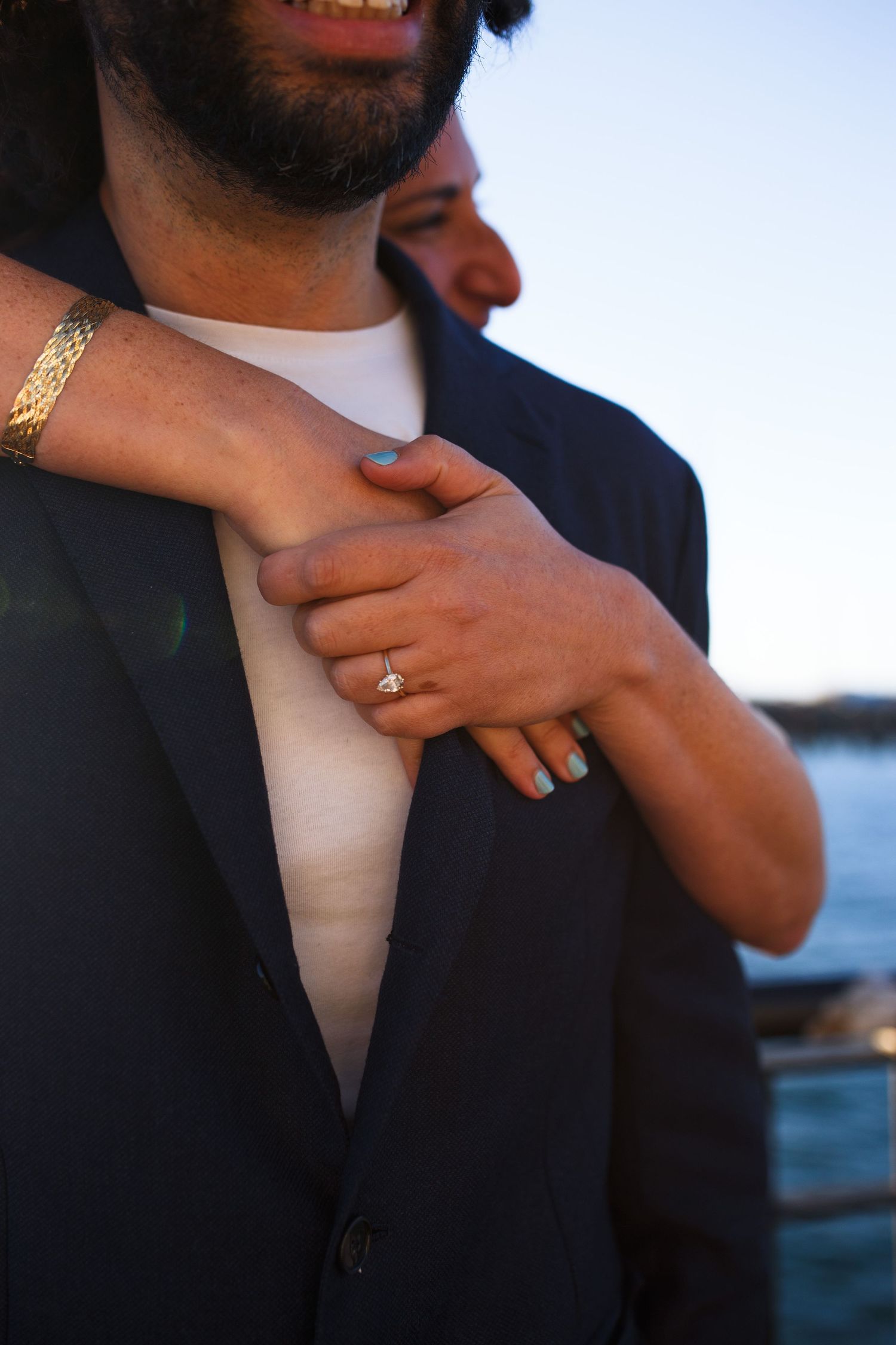 Close-up detail of a couple's hands and formal attire against an ocean backdrop at sunset.