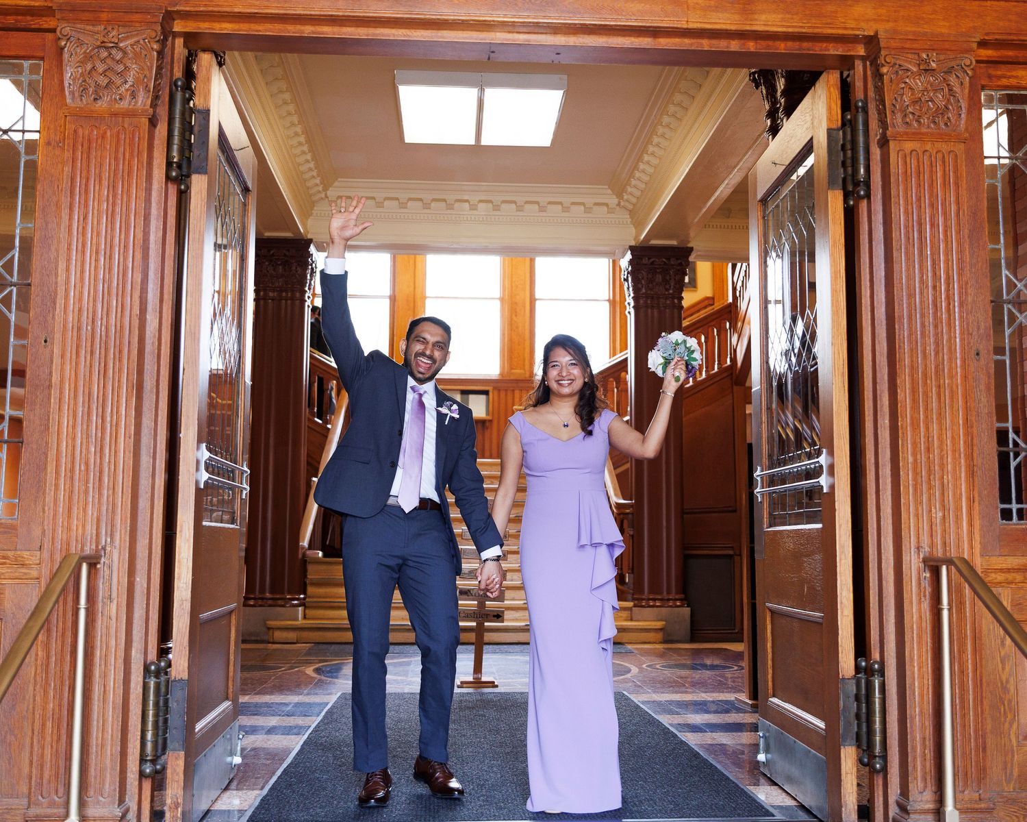 Joyful newlywed couple celebrating in elegant wood-paneled venue, bride in lavender gown holding bouquet, groom in navy suit.