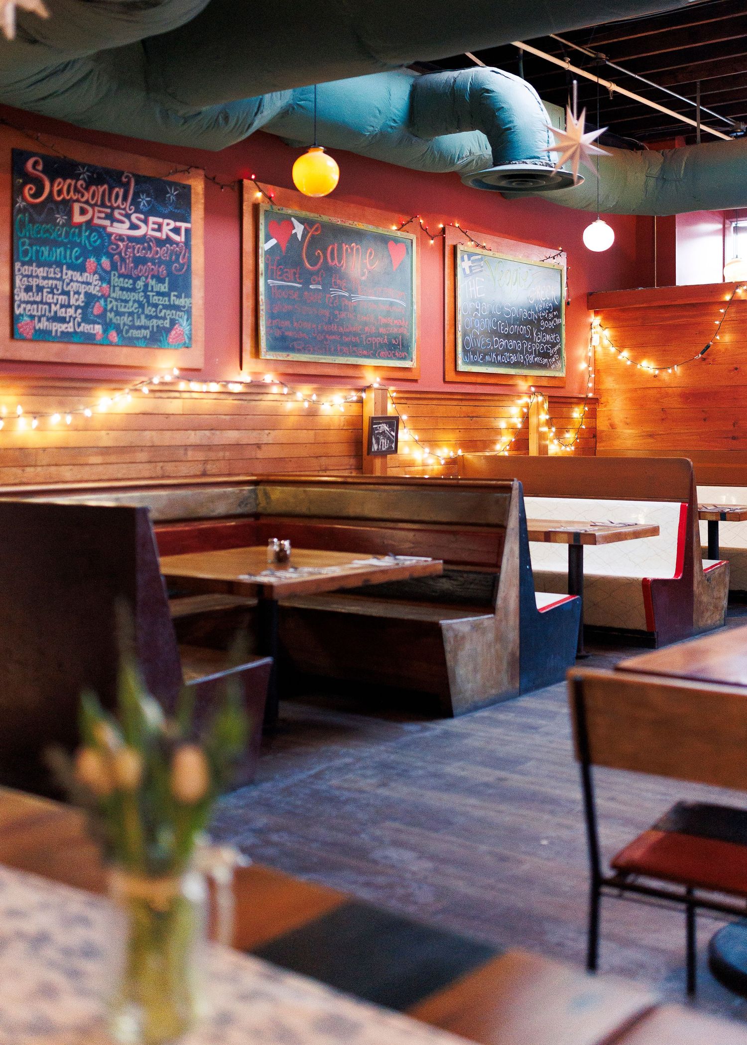 A rustic restaurant interior with wooden bench seating and chalkboard menus illuminated by warm string lights.