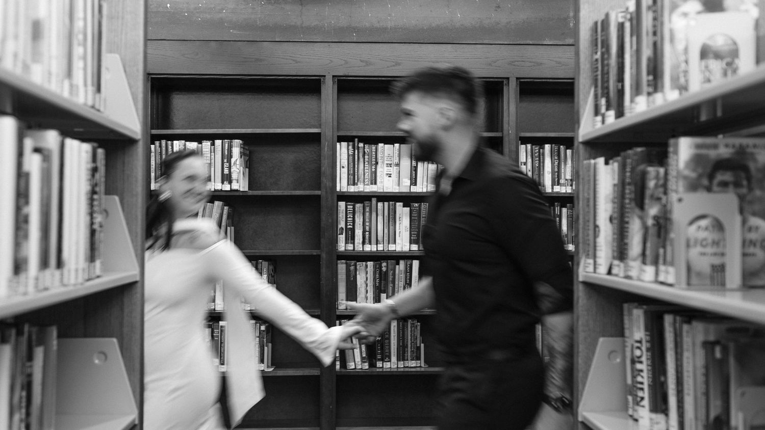 Black and white photograph showing a library interior with tall bookshelves and people browsing the aisles at Brookline Public Library.