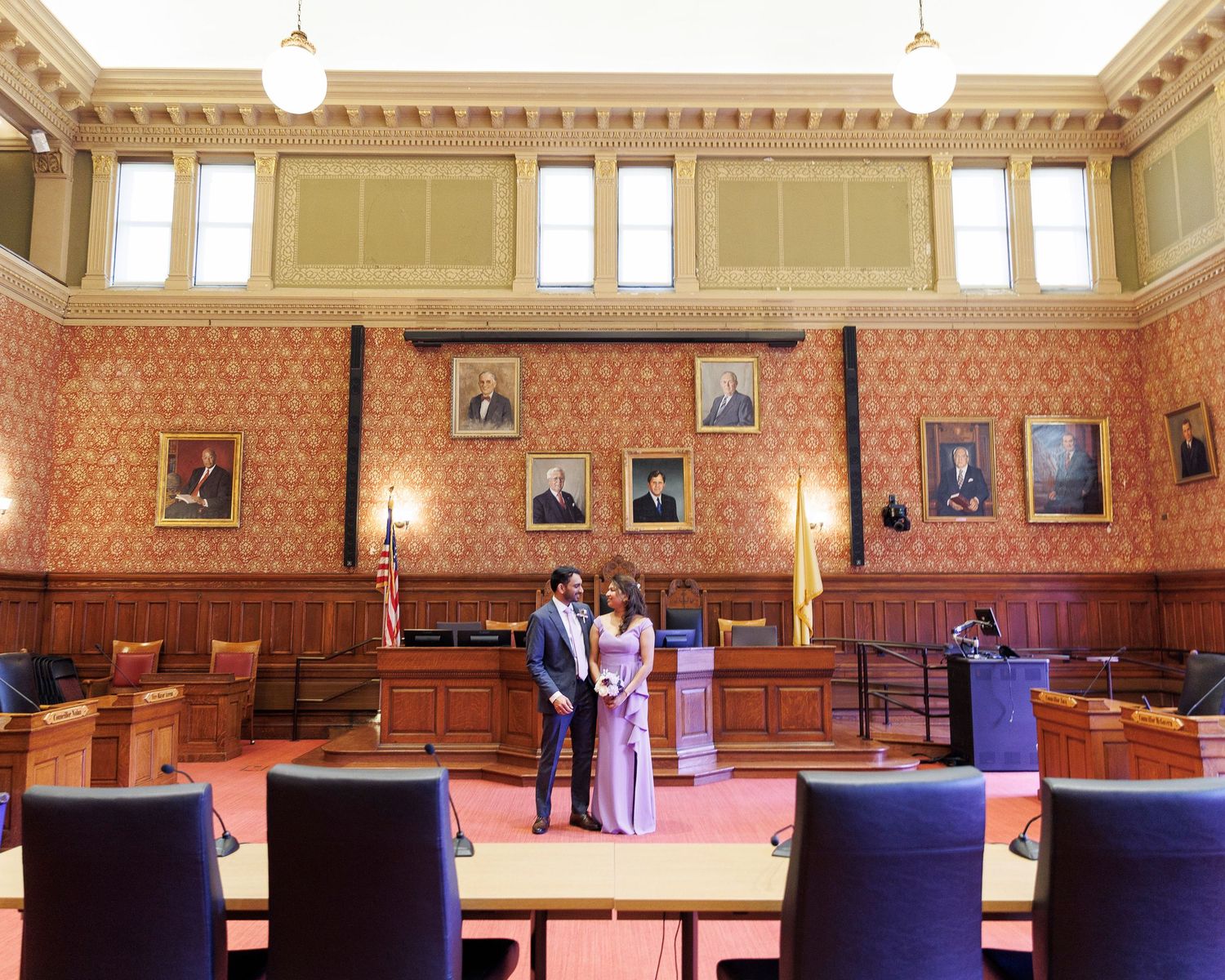 Couple standing in an ornate courtroom with red walls, portrait paintings, and purple chairs during a wedding ceremony.