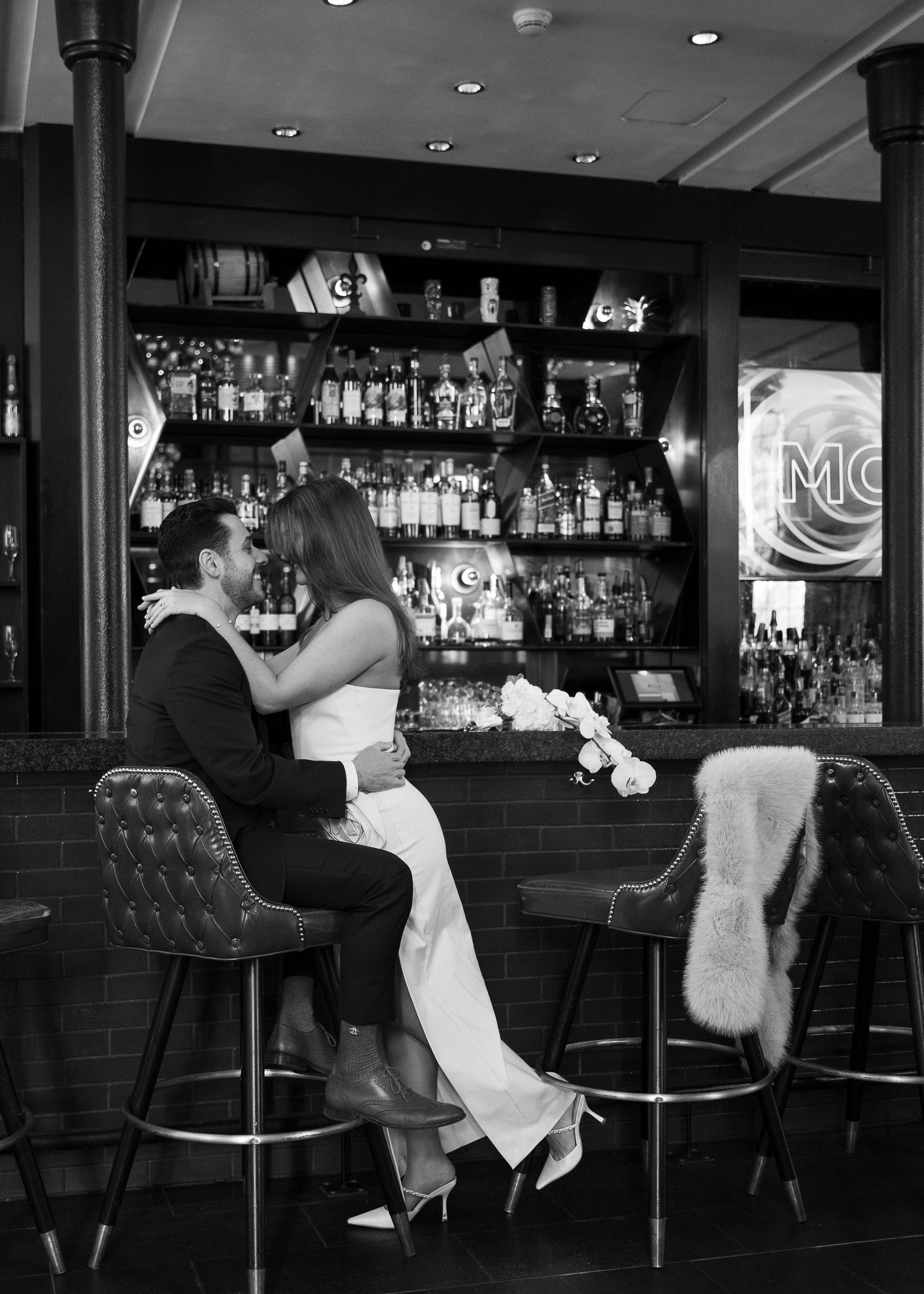 Black and white photo of a couple embracing romantically at a bar, sitting on stools with liquor bottles in background.