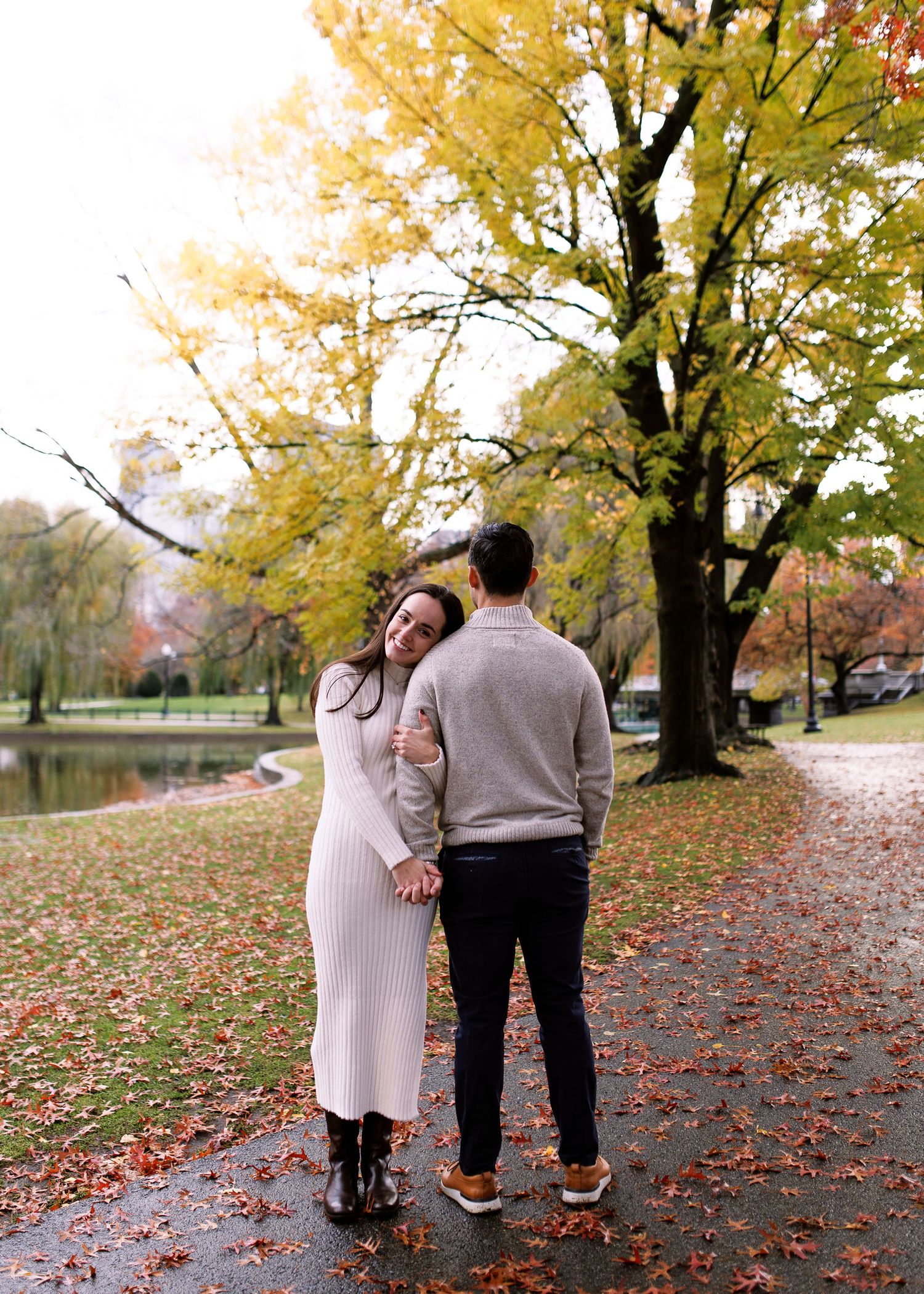 A couple walks together on a leaf-covered path beneath golden autumn trees in a park setting.