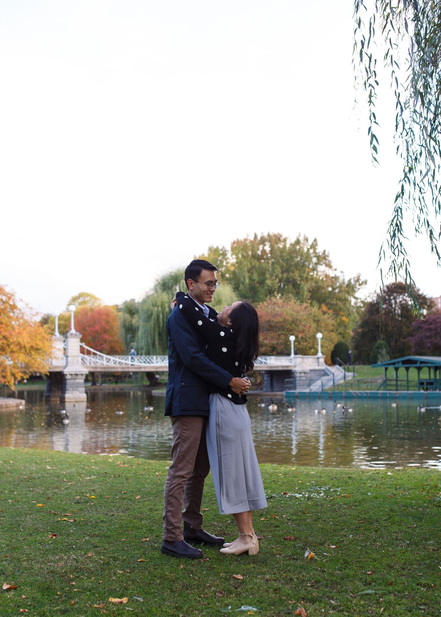Two people share a romantic moment near a pond with autumn trees and a decorative fountain in the background.