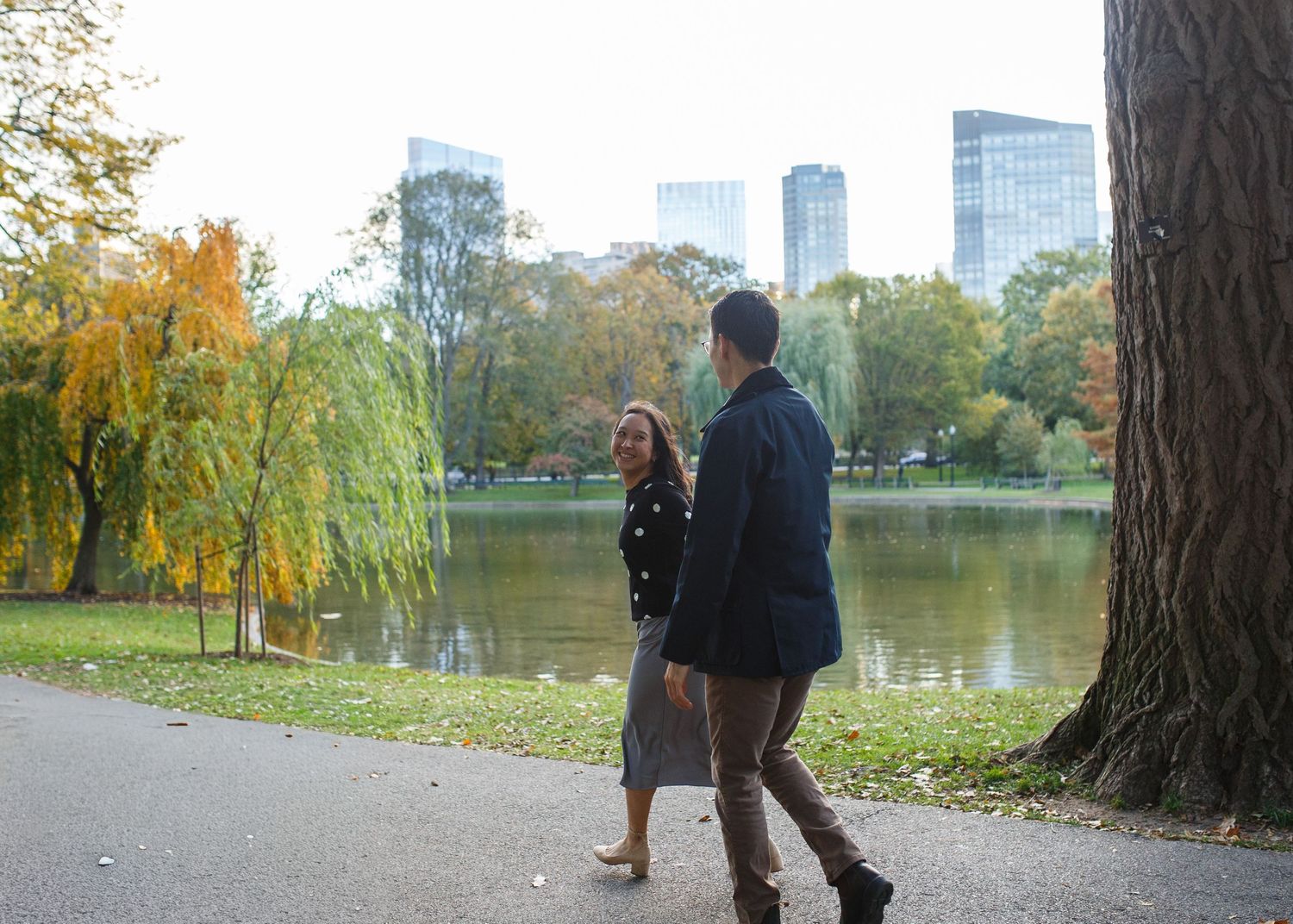 Two people stroll along a path in Central Park during autumn with skyscrapers and colorful fall foliage in the background.
