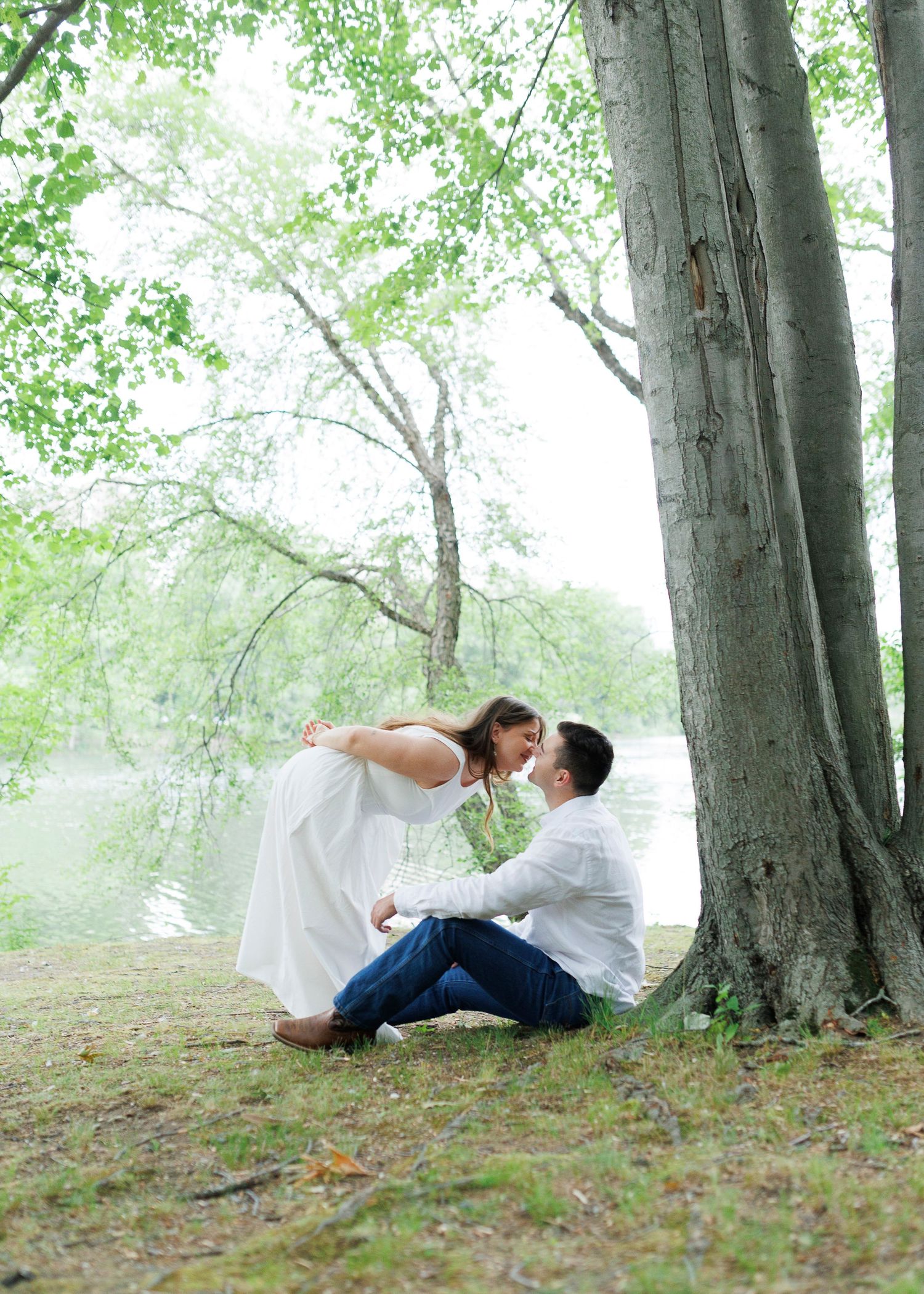 Romantic couple sharing a tender moment under a large tree by a misty lake, woman in white dress leaning toward seated man.