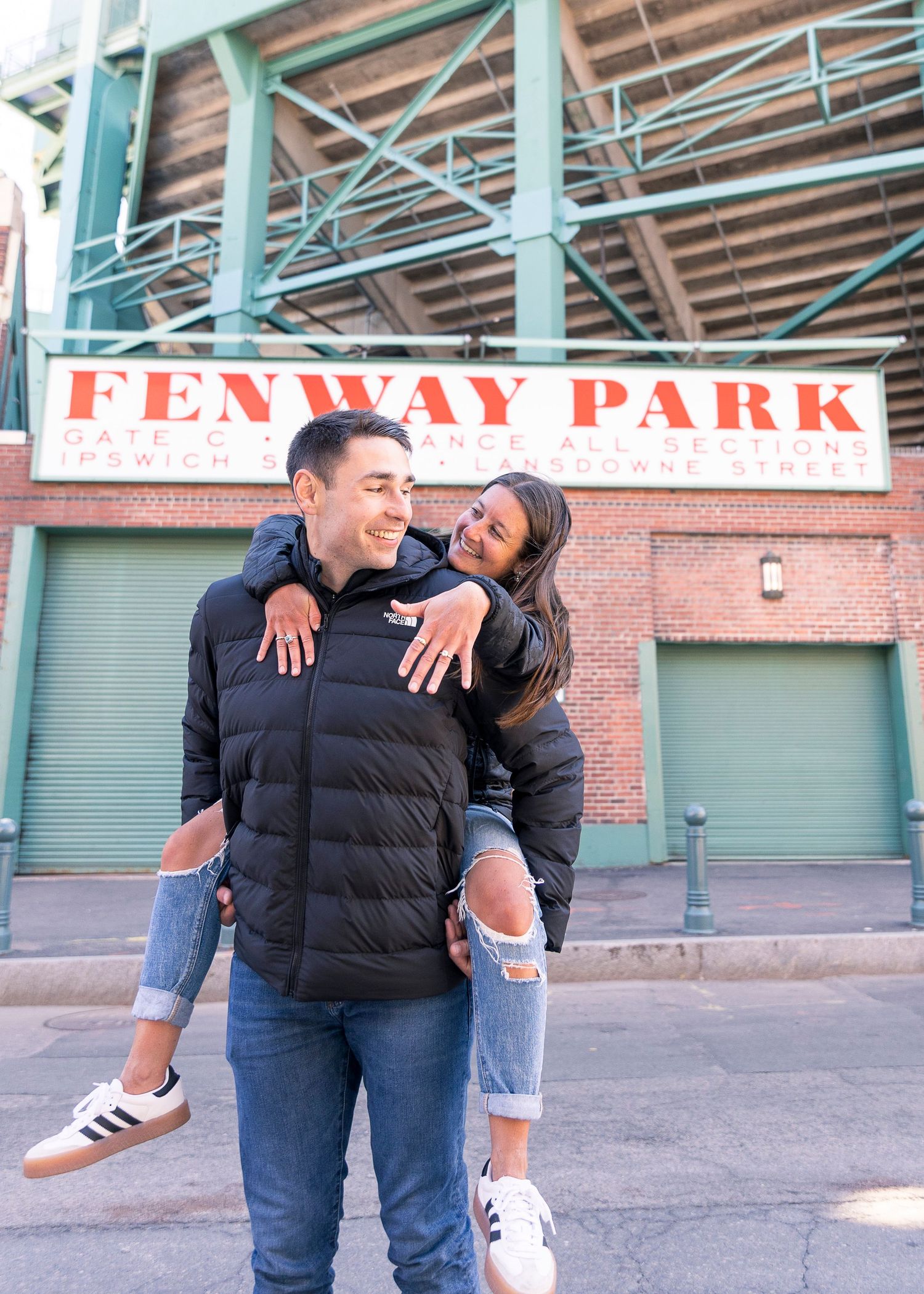 Couple posing and laughing in front of Fenway Park sign in Boston, woman on man's back wearing ripped jeans.