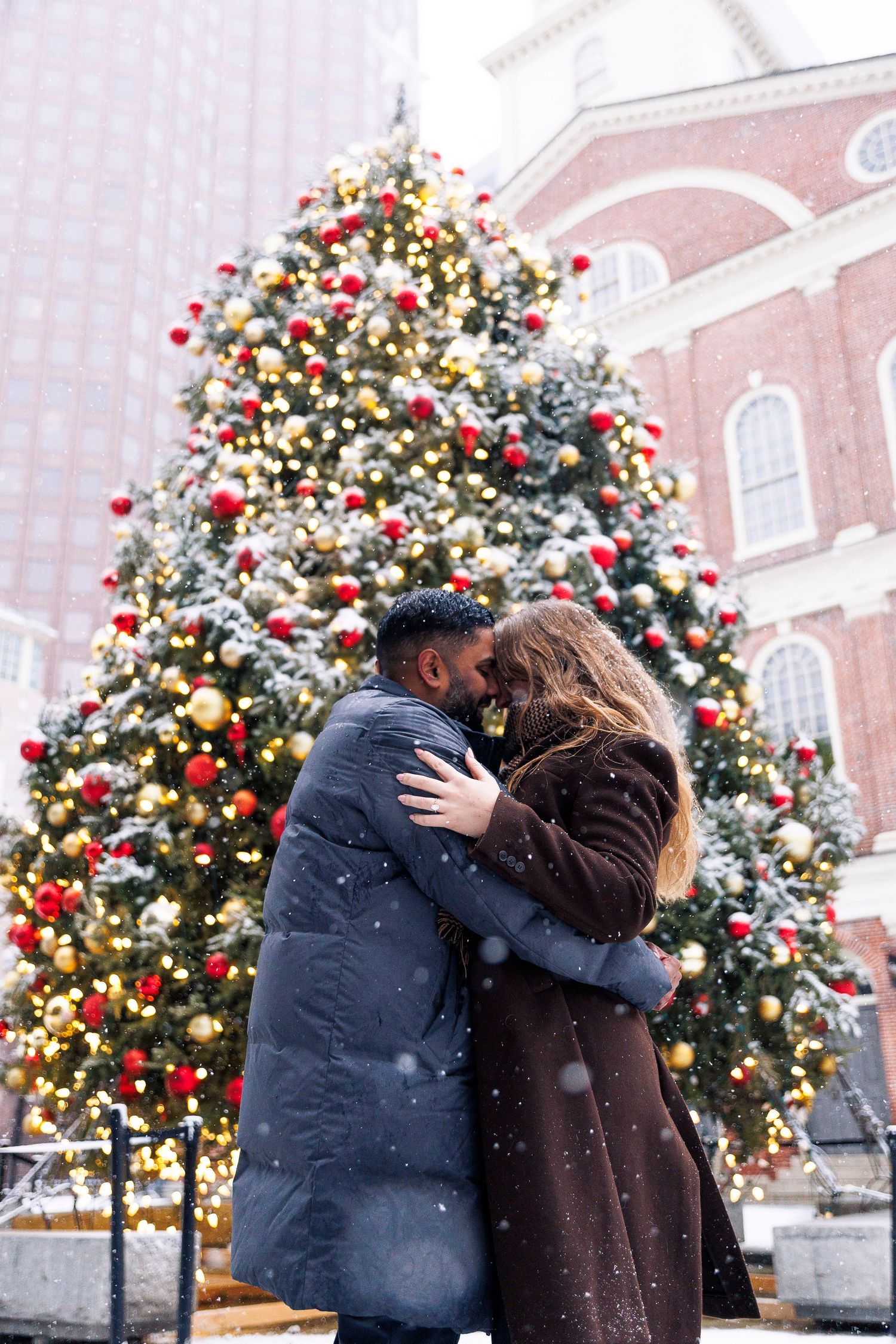 A couple shares a romantic embrace in front of a beautifully decorated Christmas tree with red and gold ornaments.