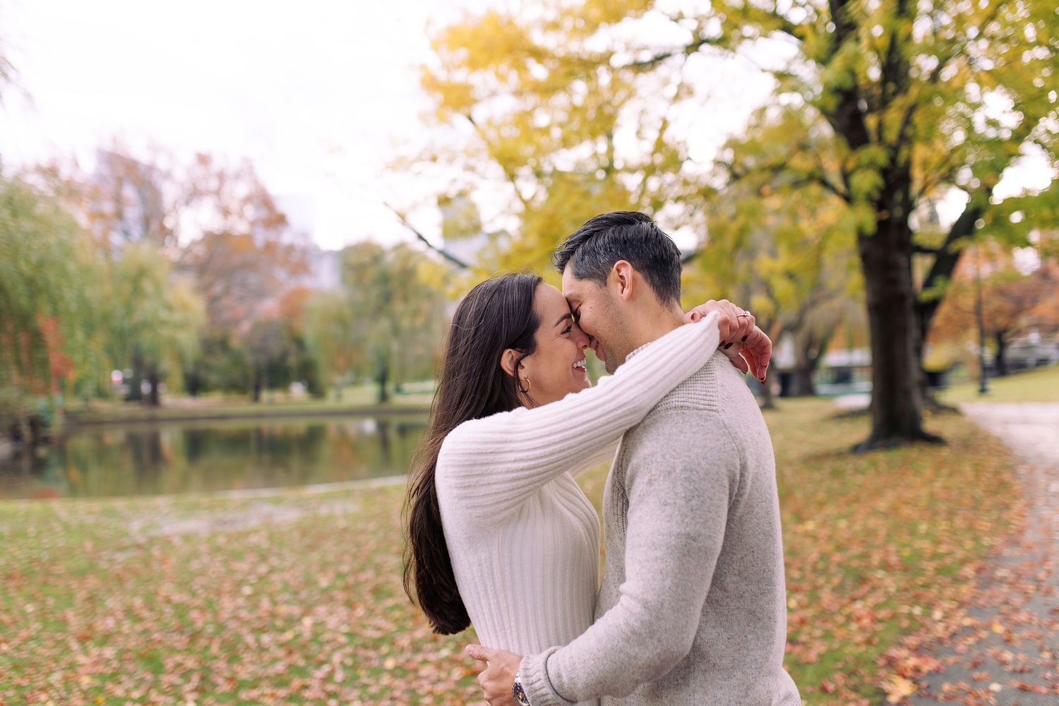 A romantic couple embraces in a park during autumn with fallen leaves and trees in the background.