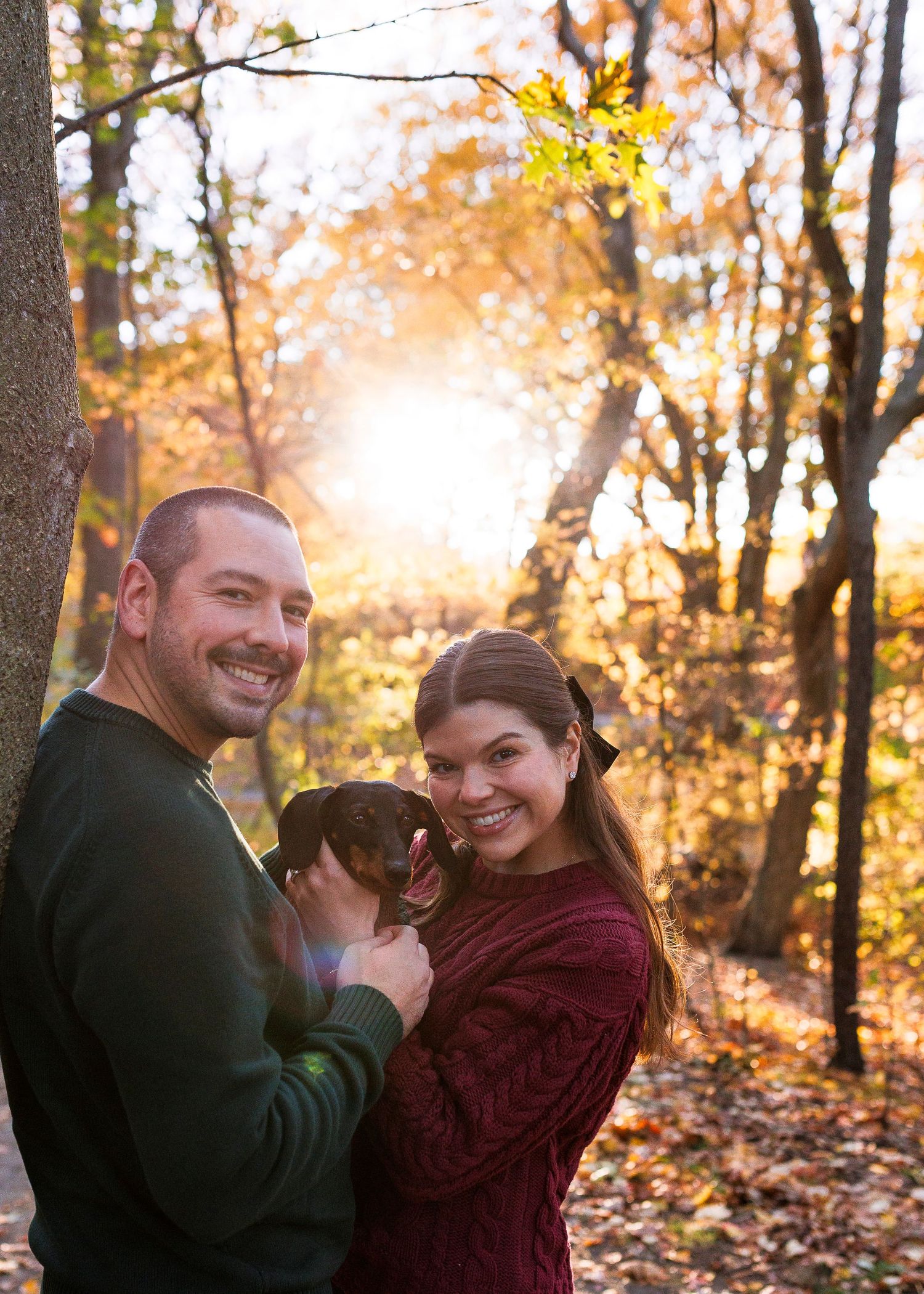 A couple and their dog share joyful moments in a sunlit autumn forest with golden leaves falling around them.
