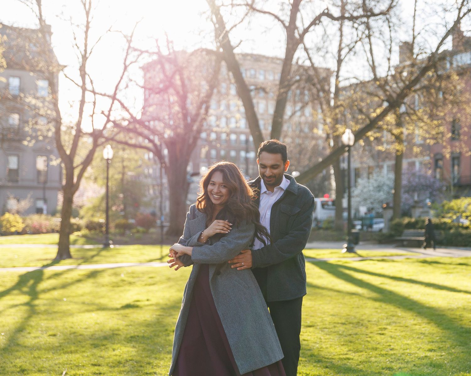 Couple embracing in a sunlit park with blooming trees and green grass in the background.