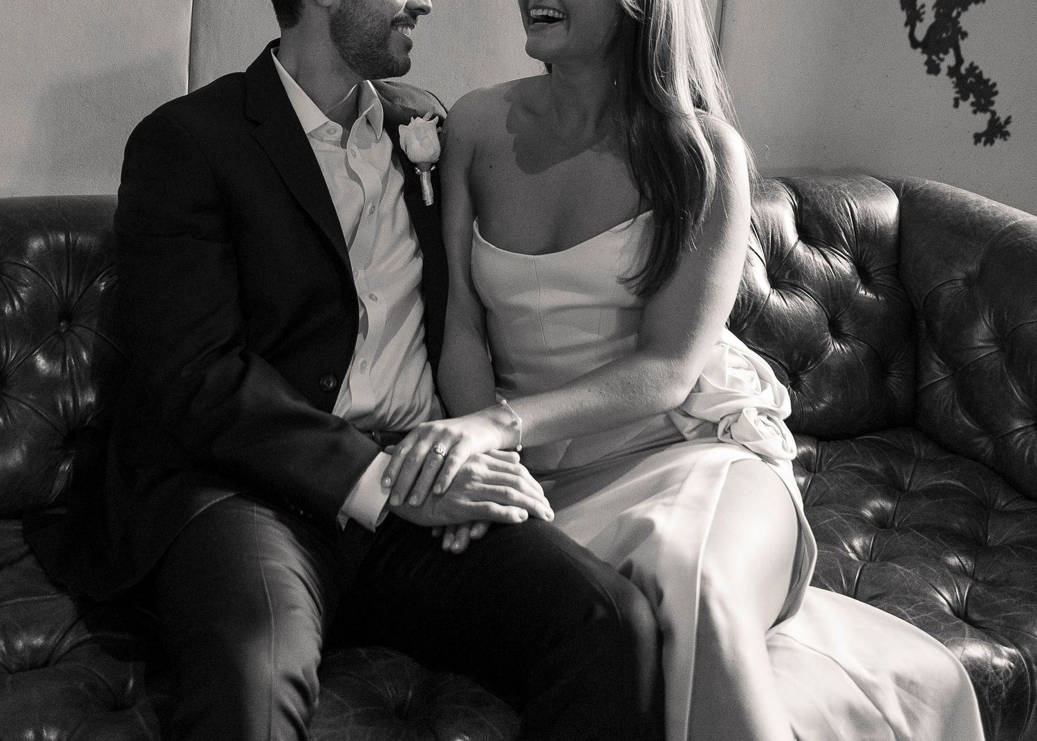 Black and white wedding photo of smiling bride and groom sitting together on a tufted leather sofa.