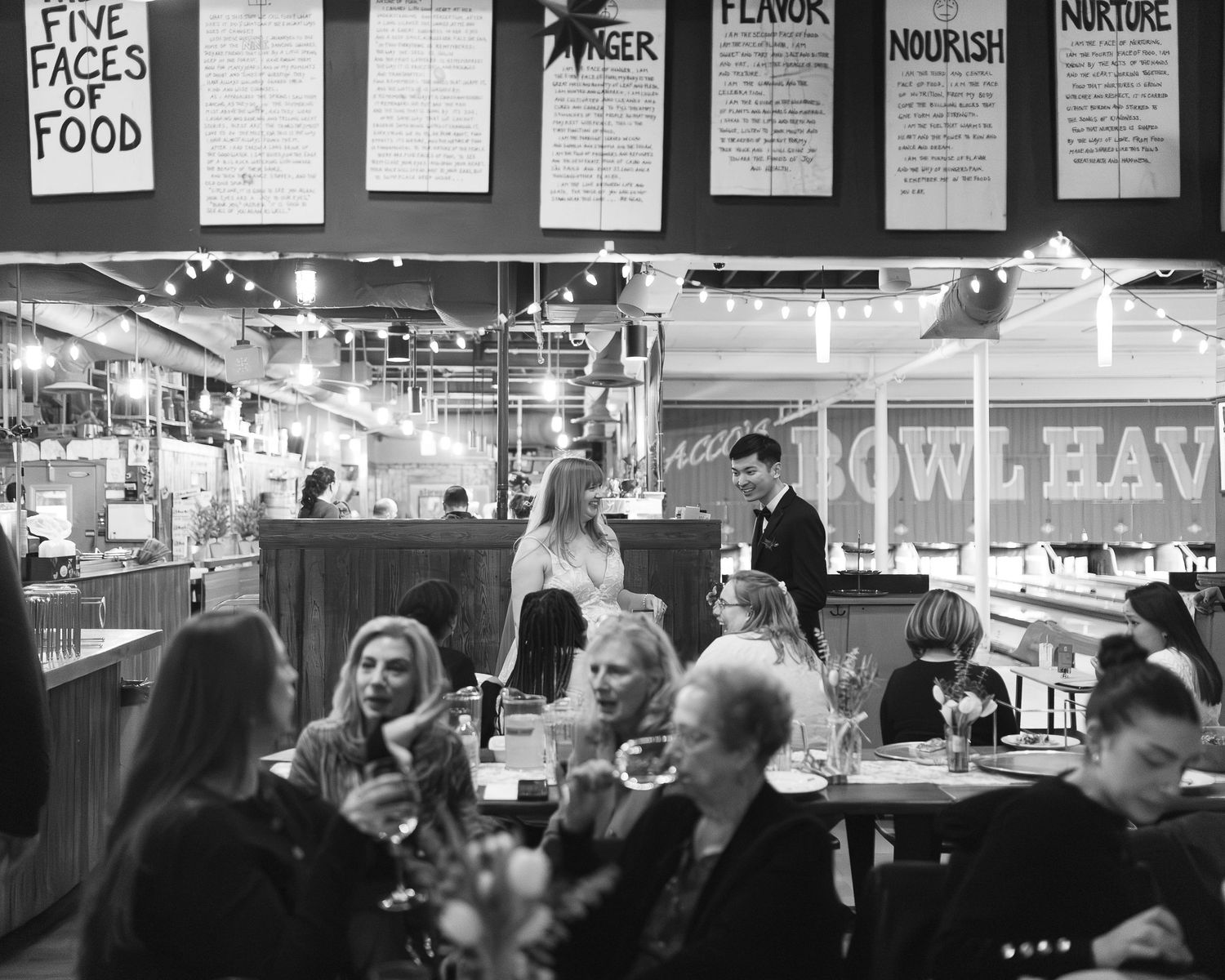 A busy restaurant interior with customers dining at tables under menu boards in black and white photography.