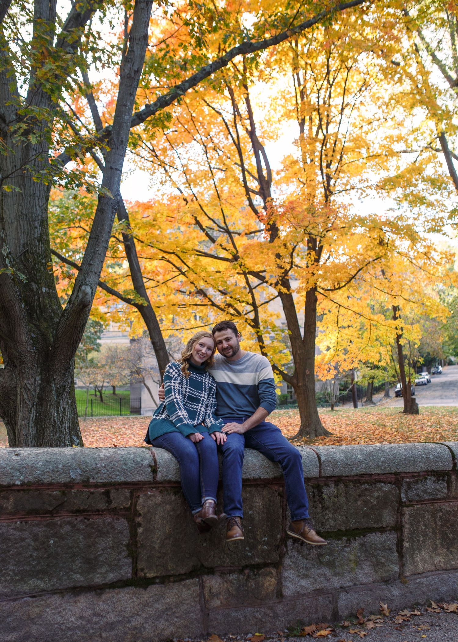 Couple sitting together on stone wall surrounded by autumn trees with vibrant yellow and orange foliage.