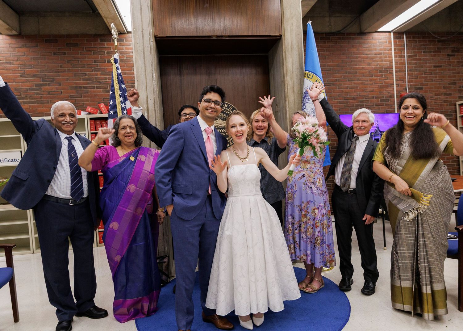 Joyful wedding party celebrating in a courthouse, with bride in white dress and guests in colorful Indian attire.