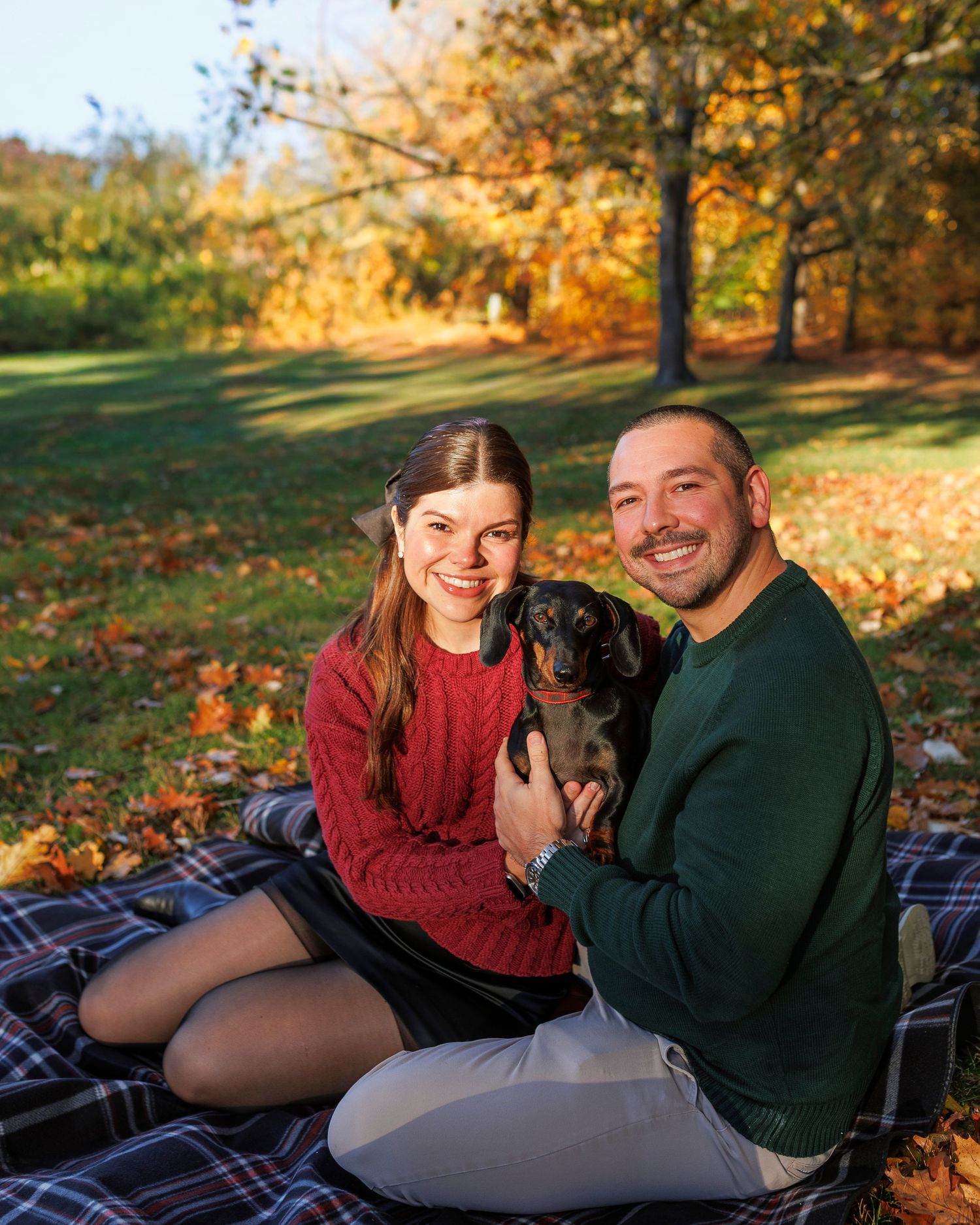 A couple and their chocolate lab puppy sit on a plaid blanket surrounded by golden autumn foliage in a park setting.