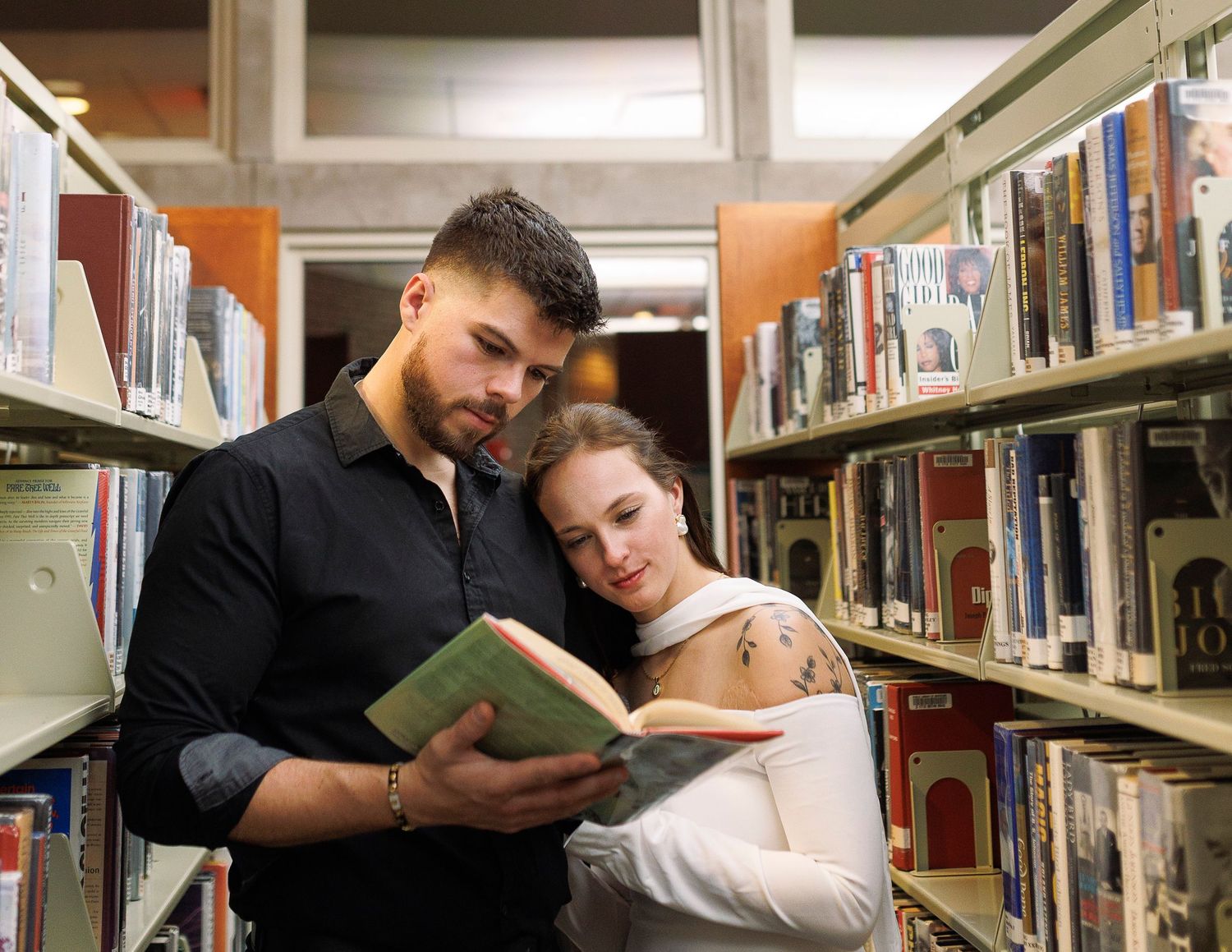Two people read together in a library while sitting close among tall bookshelves.