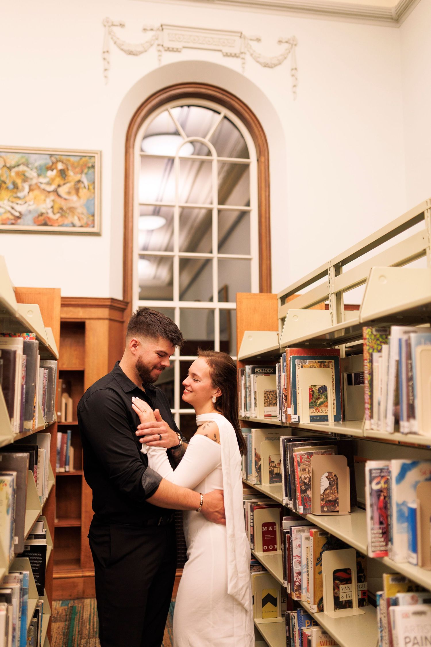 A couple shares an intimate moment between library bookshelves, with arched windows and warm lighting in the background.