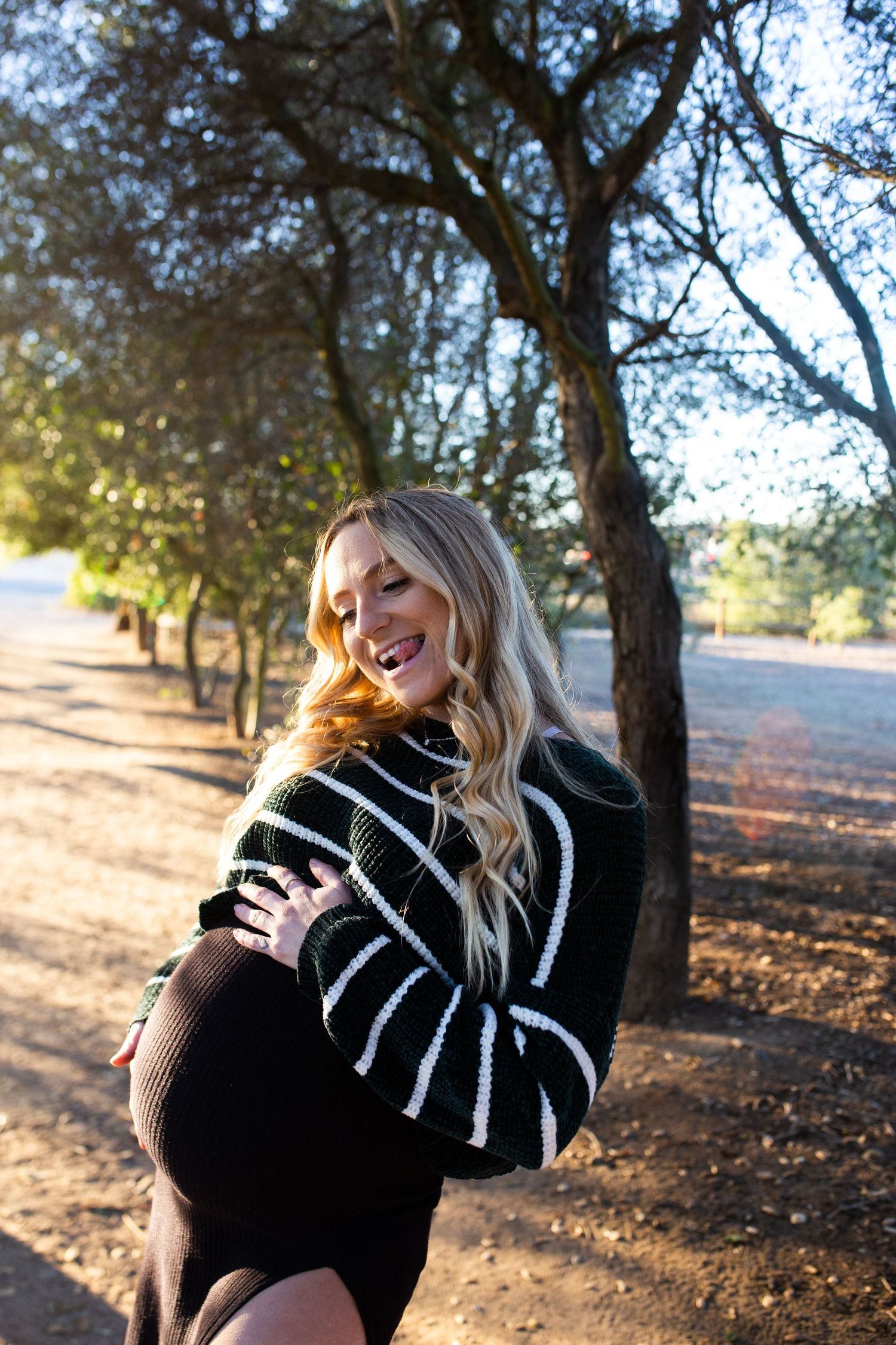 A woman in a black and white striped sweater smiles while enjoying golden hour sunlight in a park setting.