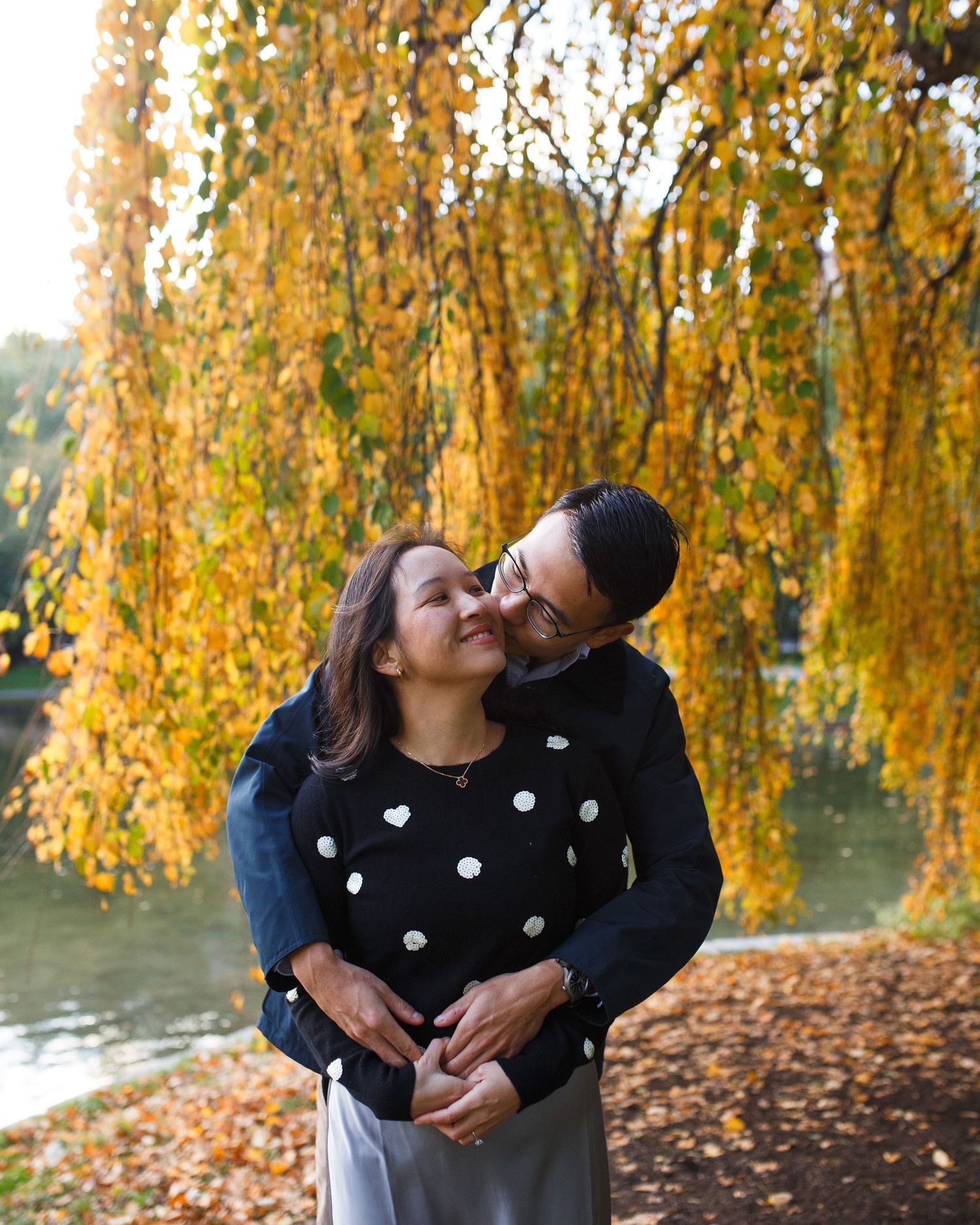A couple shares tender moments under golden autumn trees by a lake as fallen leaves scatter around them.