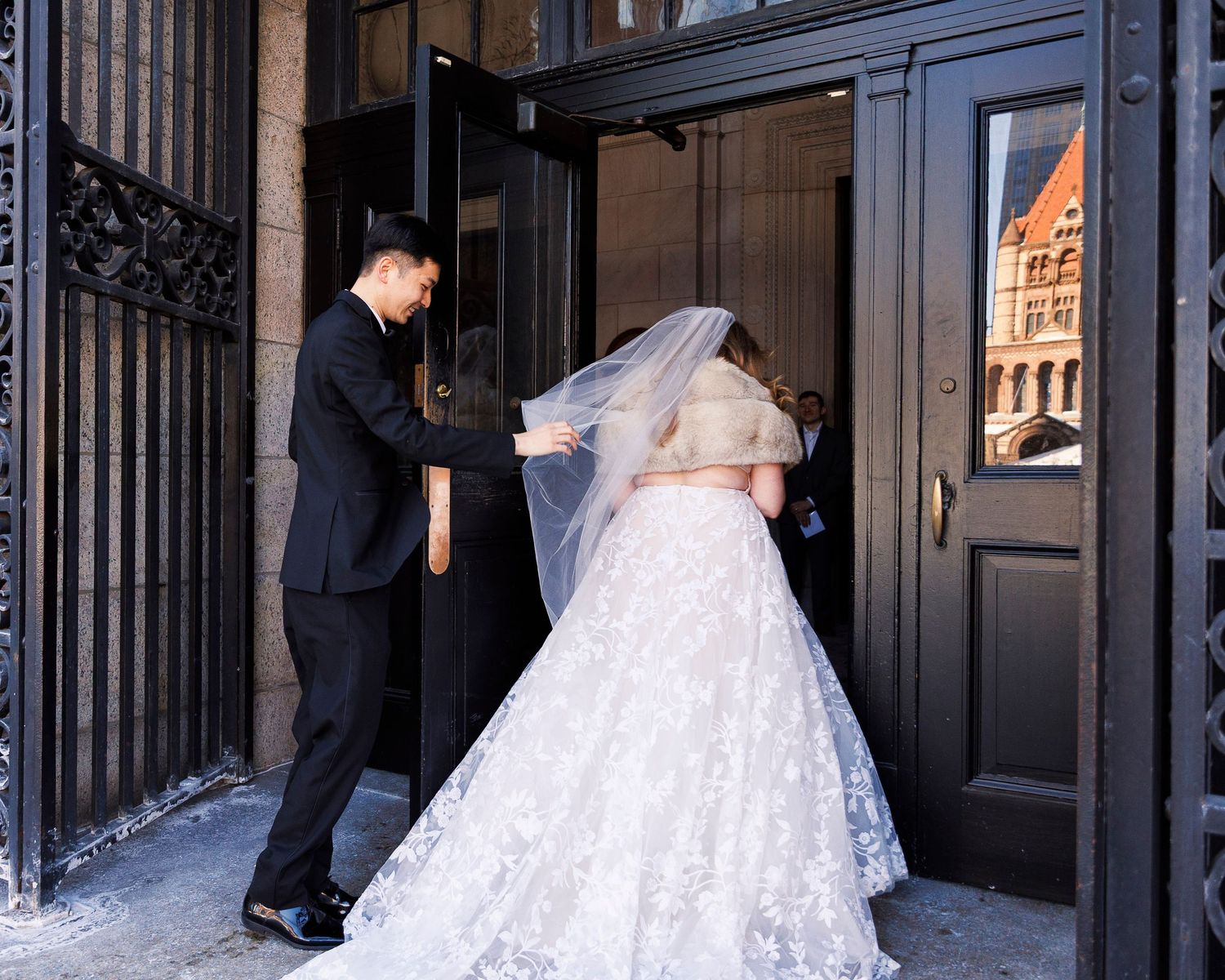 A bride in a flowing white gown with a long train walks through a doorway while someone holds the door.