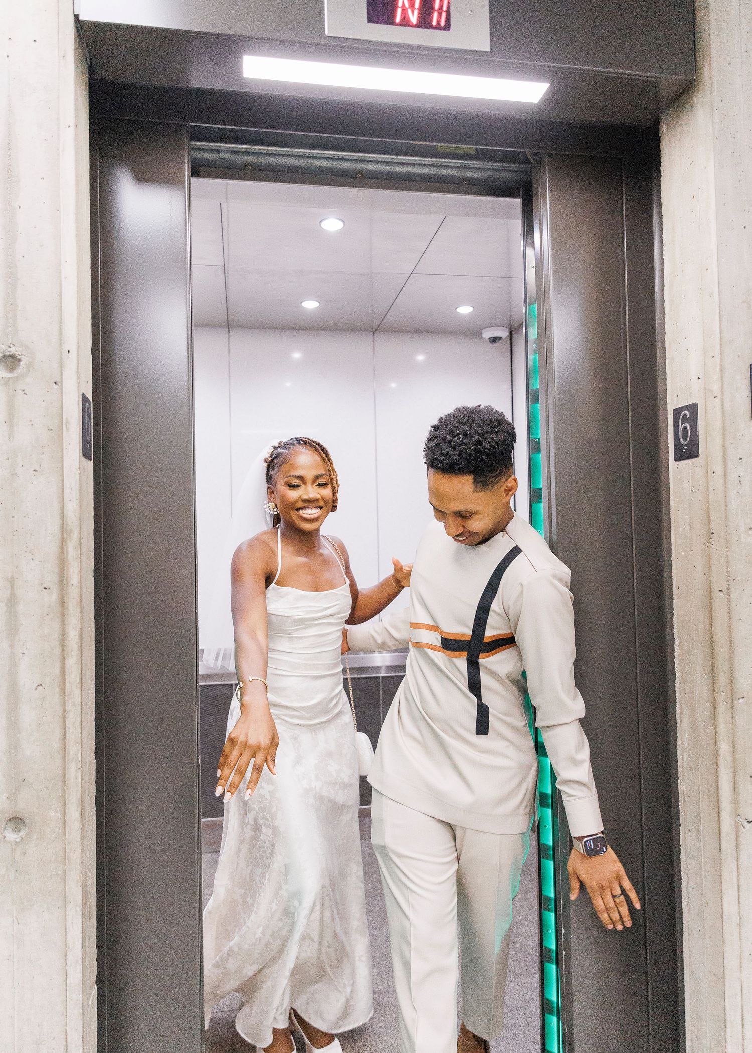 A joyful couple in white wedding attire stepping out of a modern elevator, smiling and celebrating.