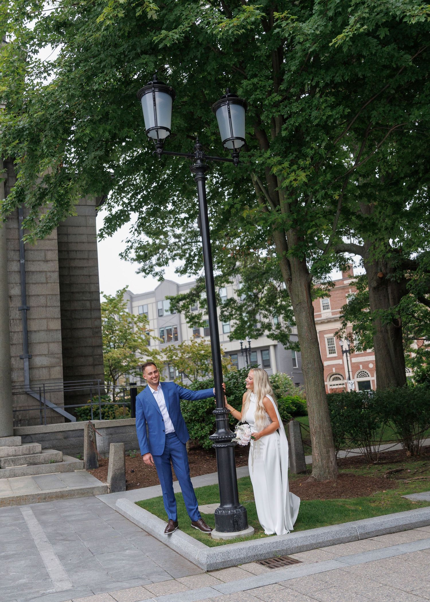 Bride and groom posing by a vintage lamp post outdoors, surrounded by lush green trees near a historic building.