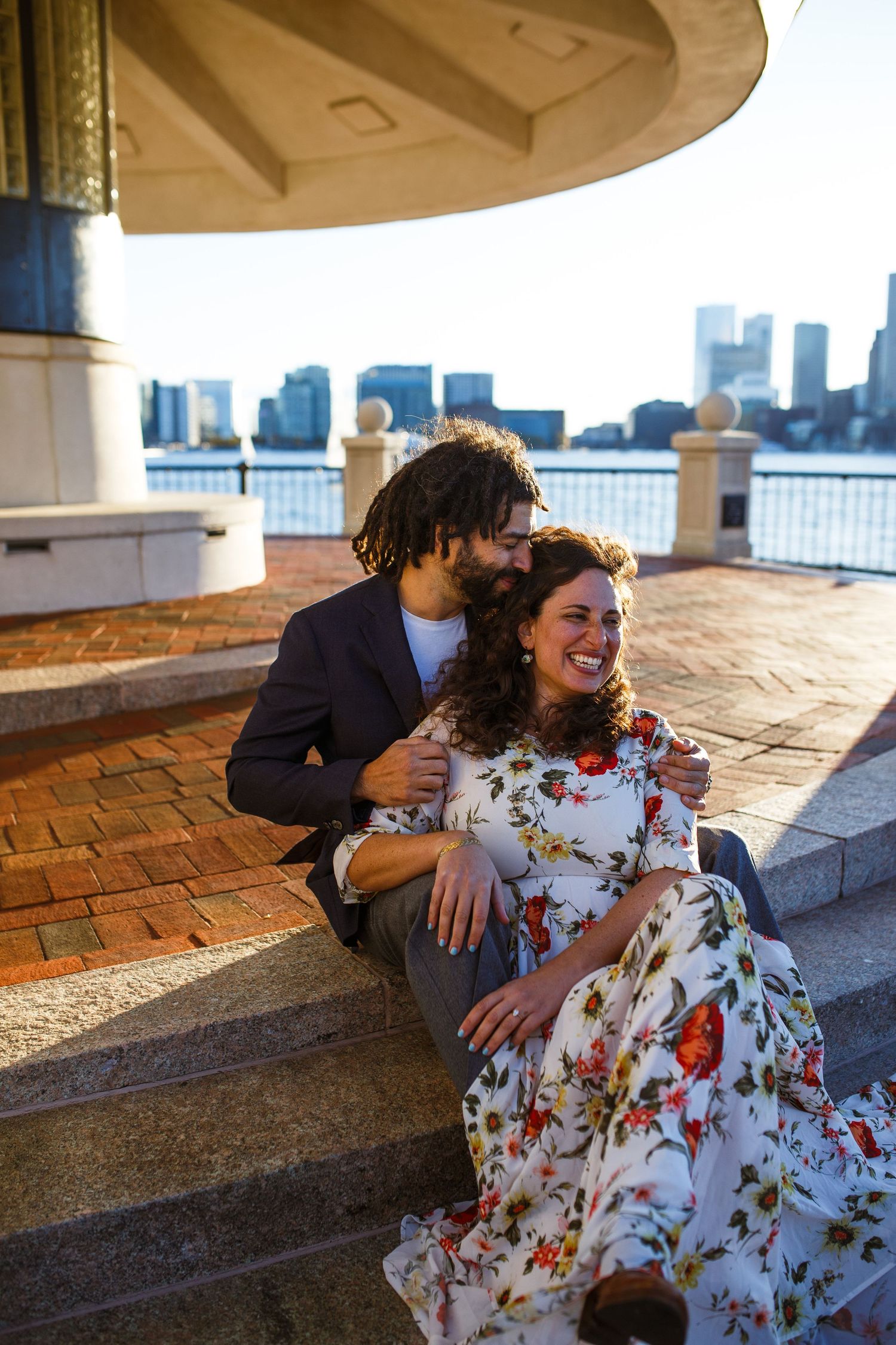 Couple laughing and embracing on brick terrace overlooking waterfront with city skyline in distance.