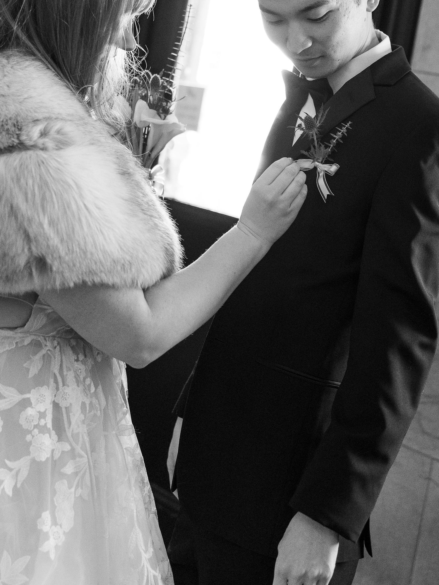 A bride in a white dress with fur trim pins a boutonniere onto a groom's black suit jacket during a wedding ceremony.