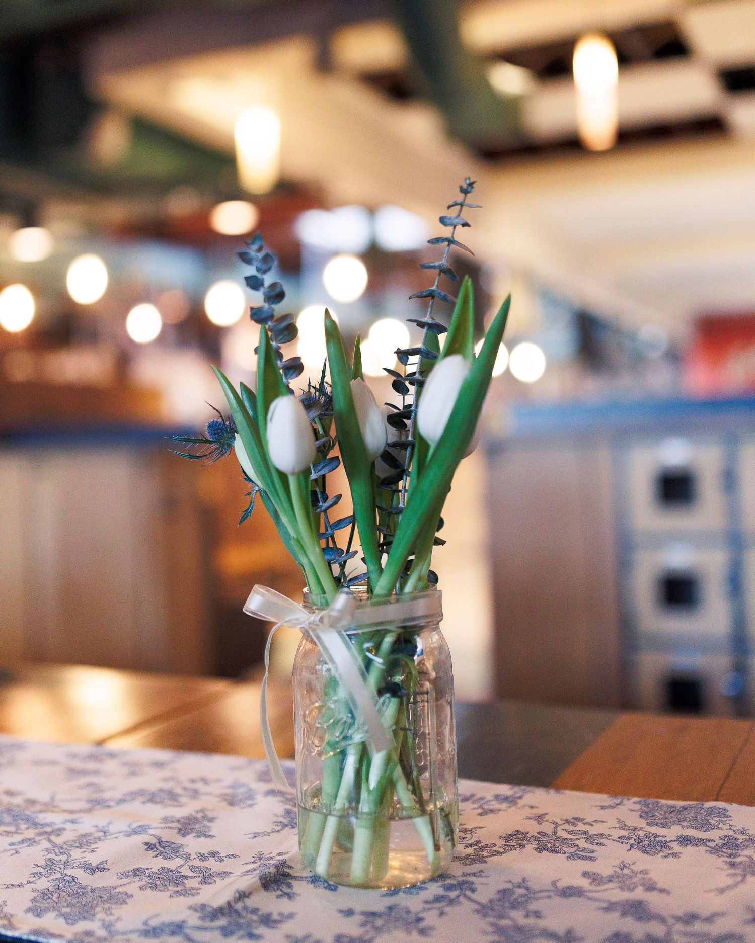 White tulips in a glass jar sit on a blue and white patterned tablecloth with soft bokeh lights in the background.