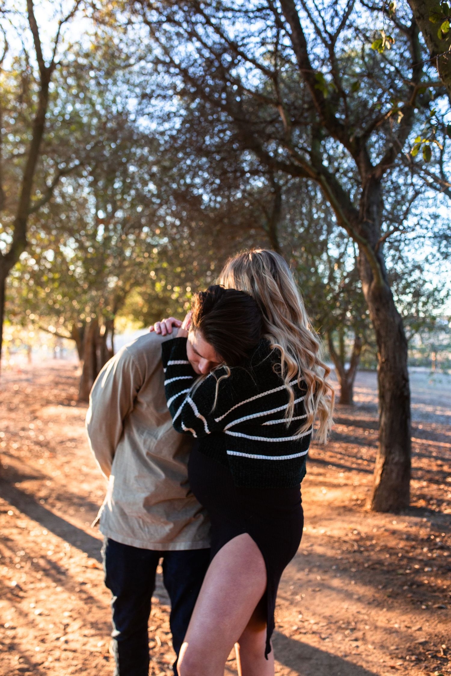 Two people share a tender embrace in a wooded area during golden hour as sunlight filters through autumn trees.