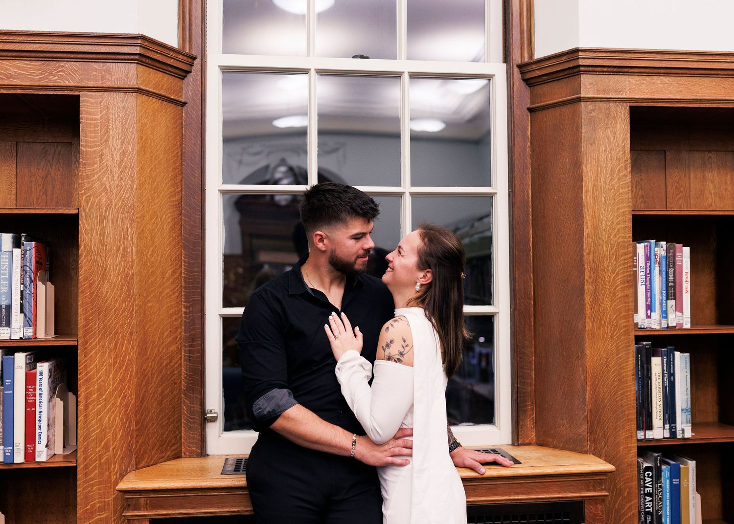 A couple shares a tender moment in a library setting with wooden bookshelves in the background.