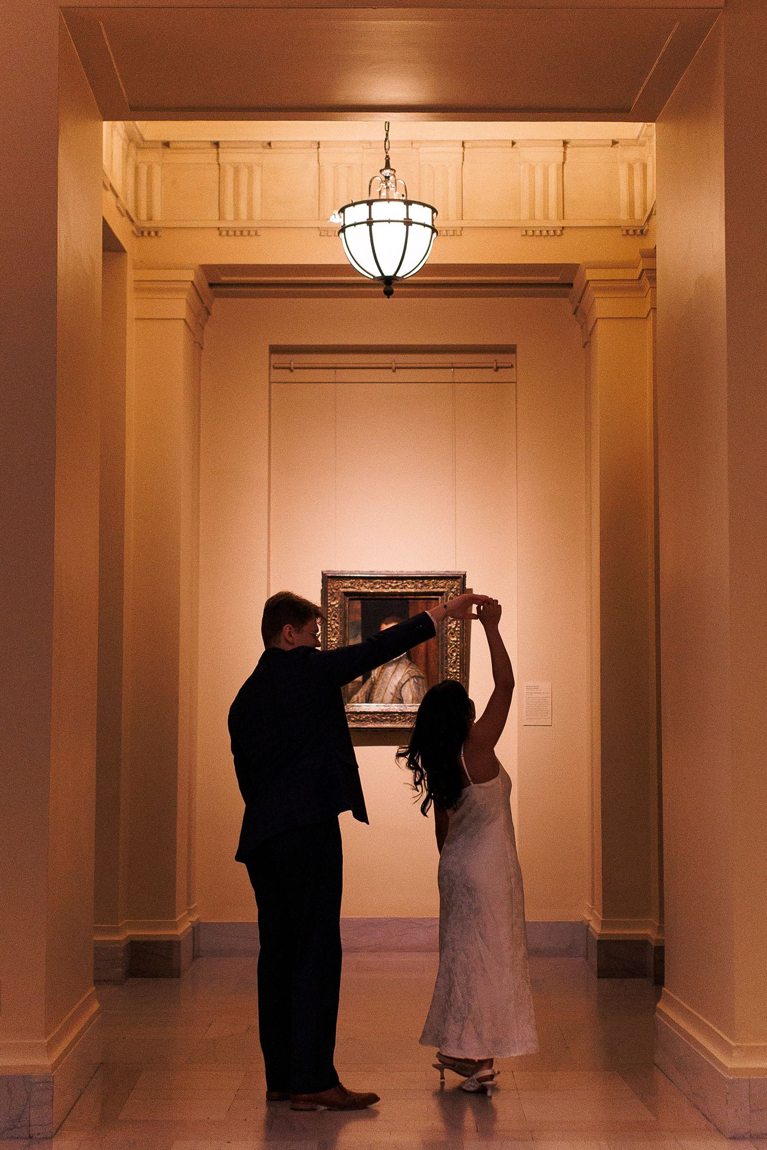 Two silhouettes dance together in an elegant hallway with classical architectural details and warm lighting.