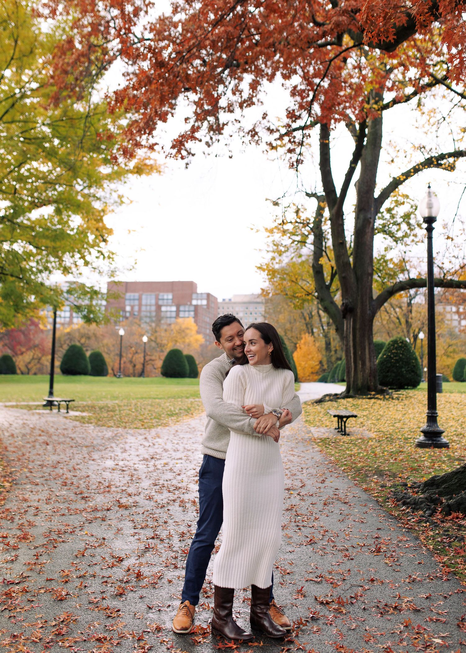 Dancing together on leaf-covered ground beneath autumn trees with brick buildings in the distance.